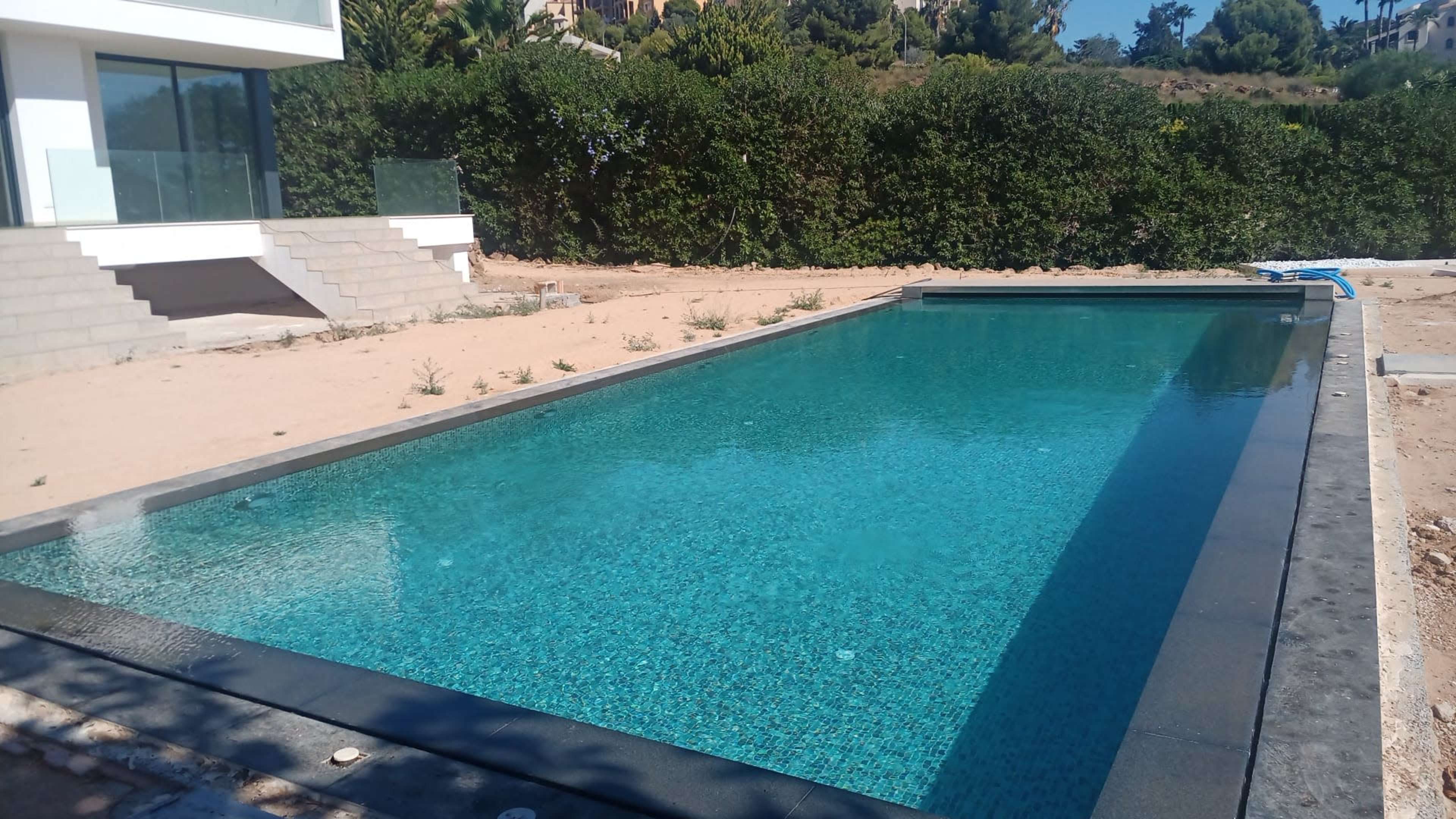 A clear swimming pool is situated beside a modern building, surrounded by bare soil and greenery.