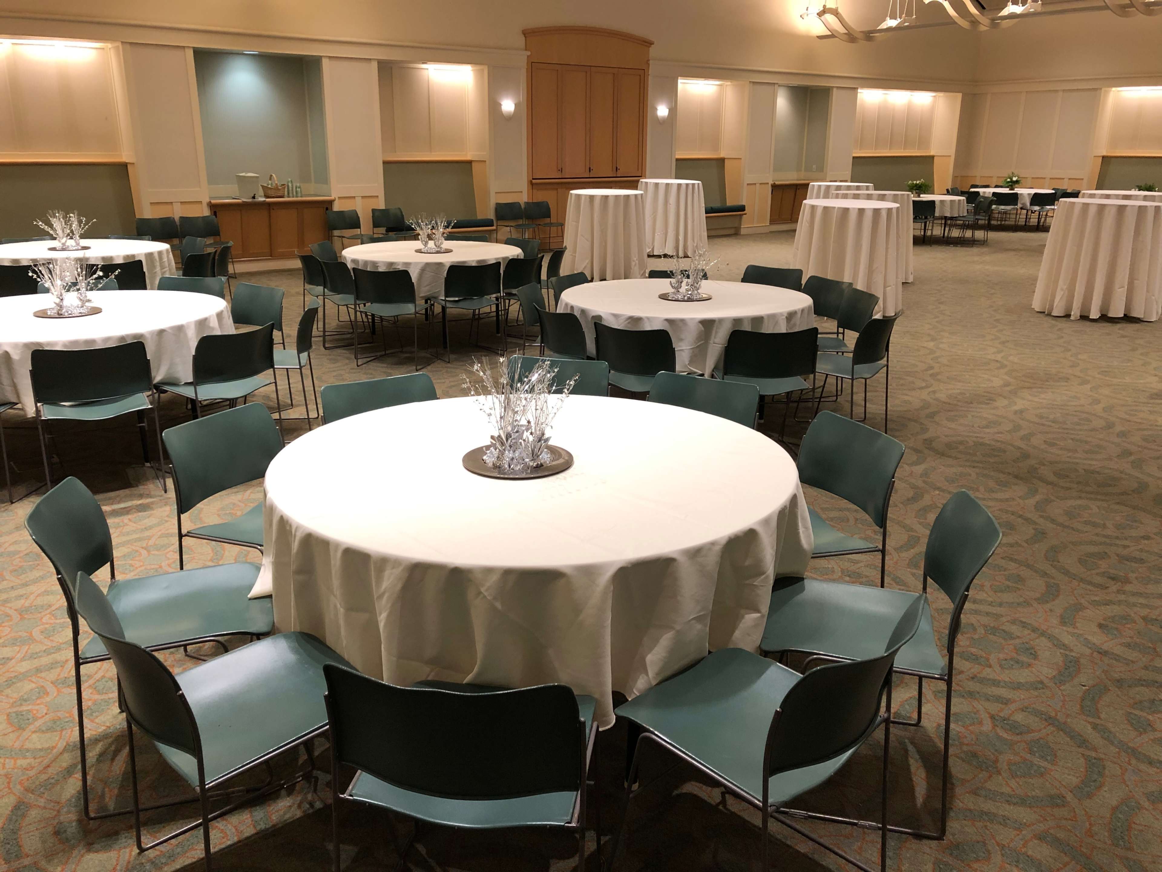A spacious room is set up with several round tables covered in white tablecloths and surrounded by green chairs.