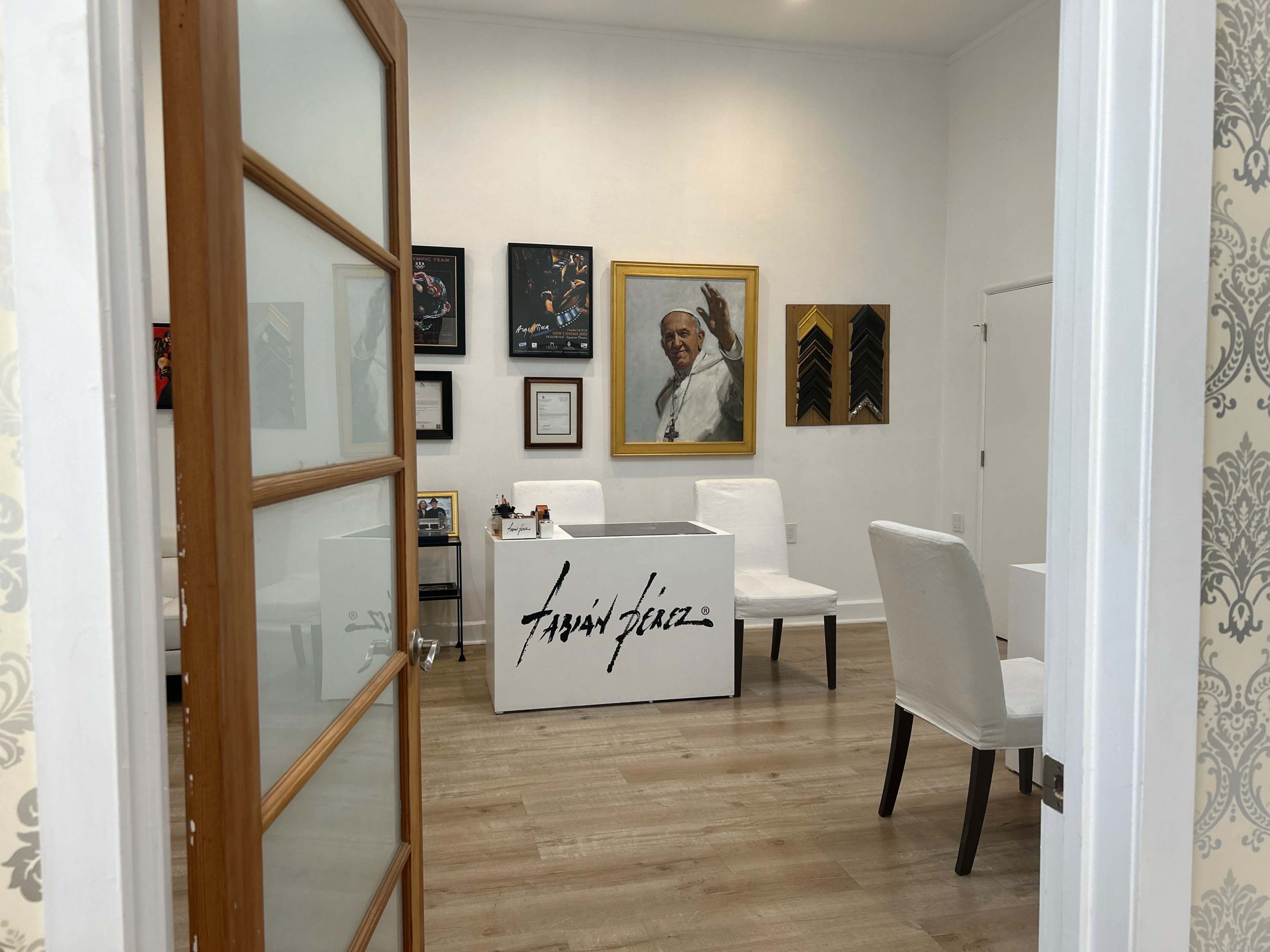 The image shows an office space with a white table featuring the name "Fabian Perez," surrounded by four white chairs and decorated with framed artworks on the walls.