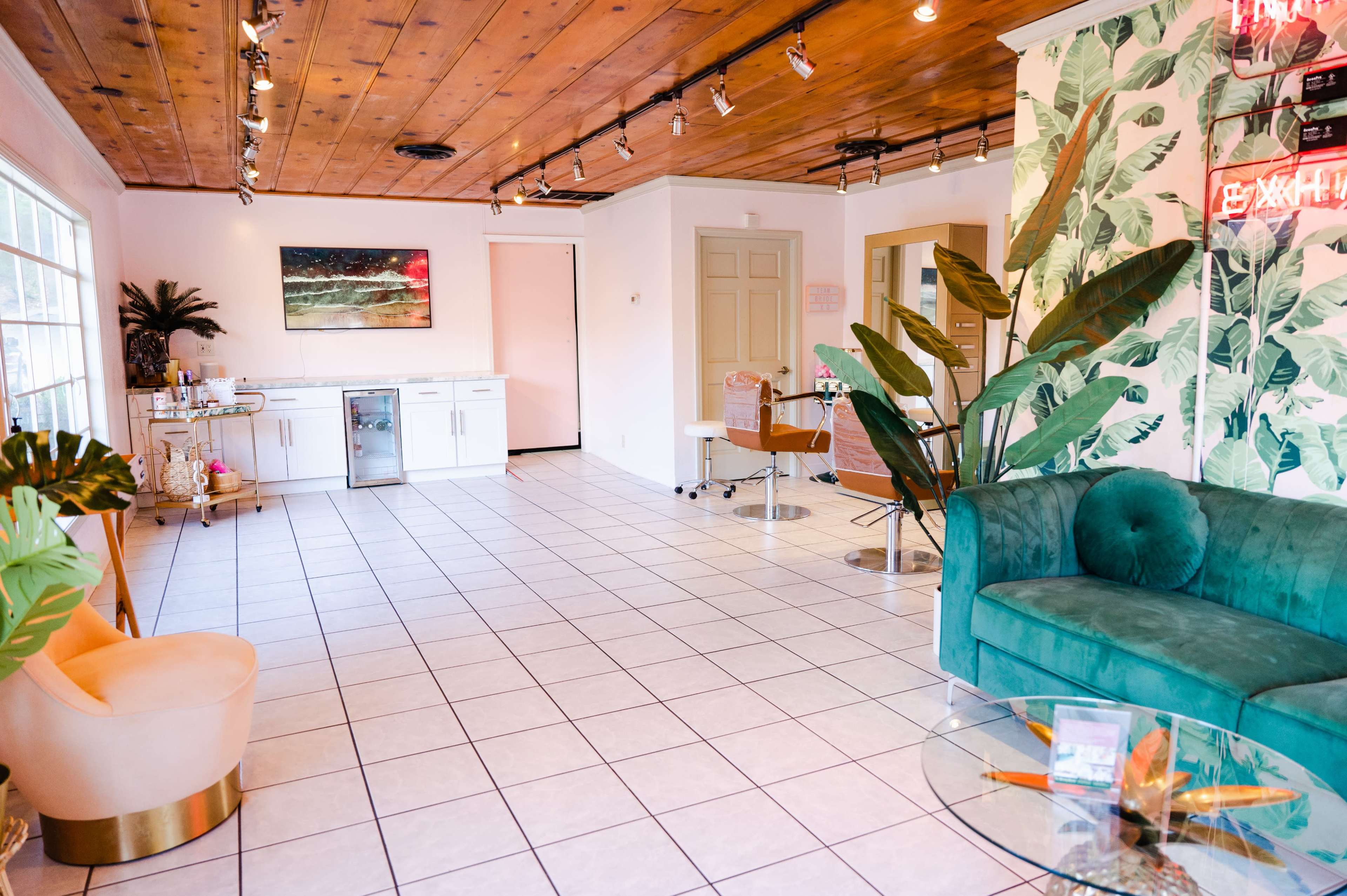 A salon interior with tiled flooring, a green sofa, a coffee table, and a kitchenette area against a backdrop of tropical-themed wallpaper.