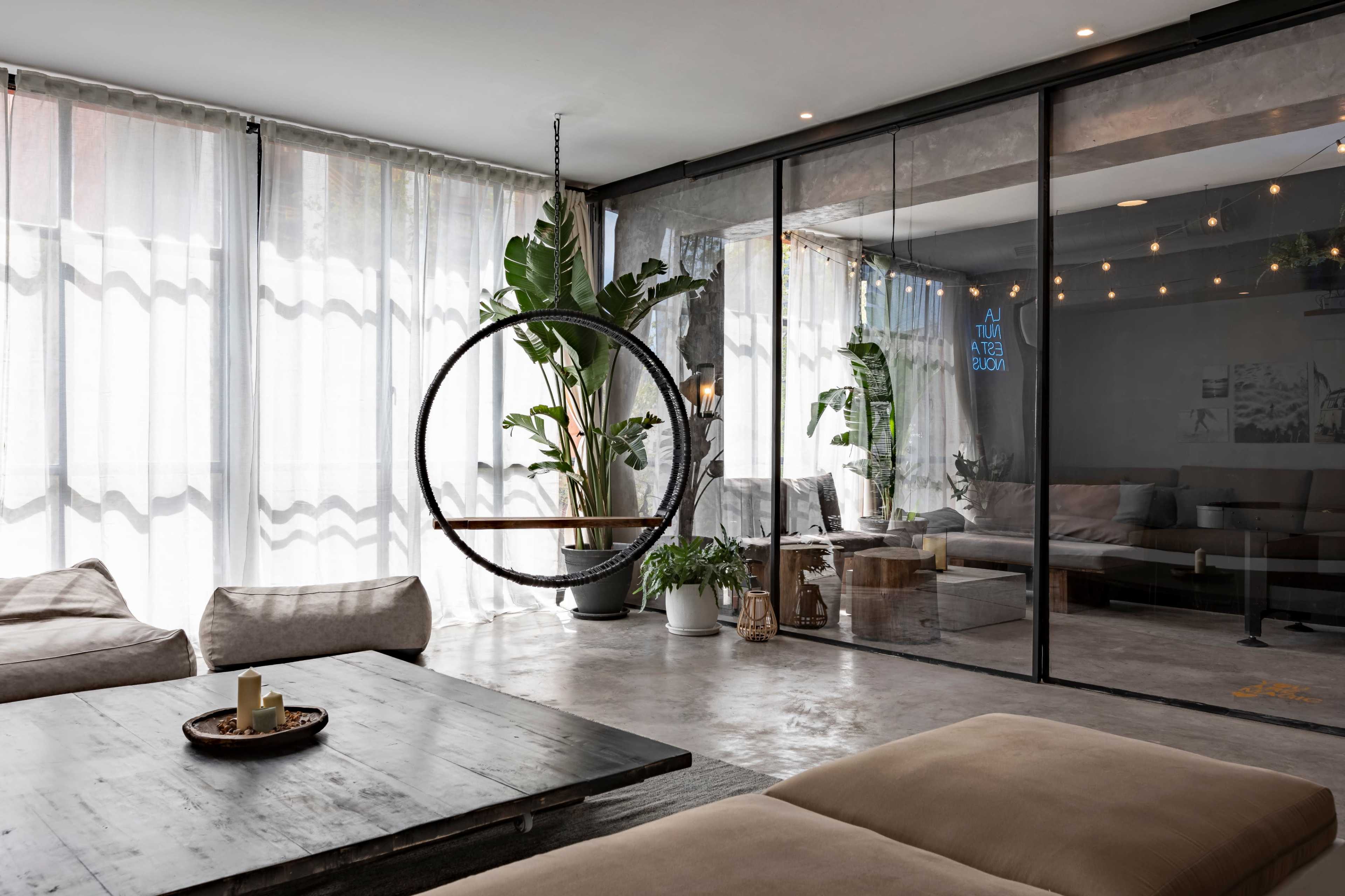 A modern living room features a circular hanging chair, large windows with sheer curtains, and a glass wall separating it from a cozy seating area.