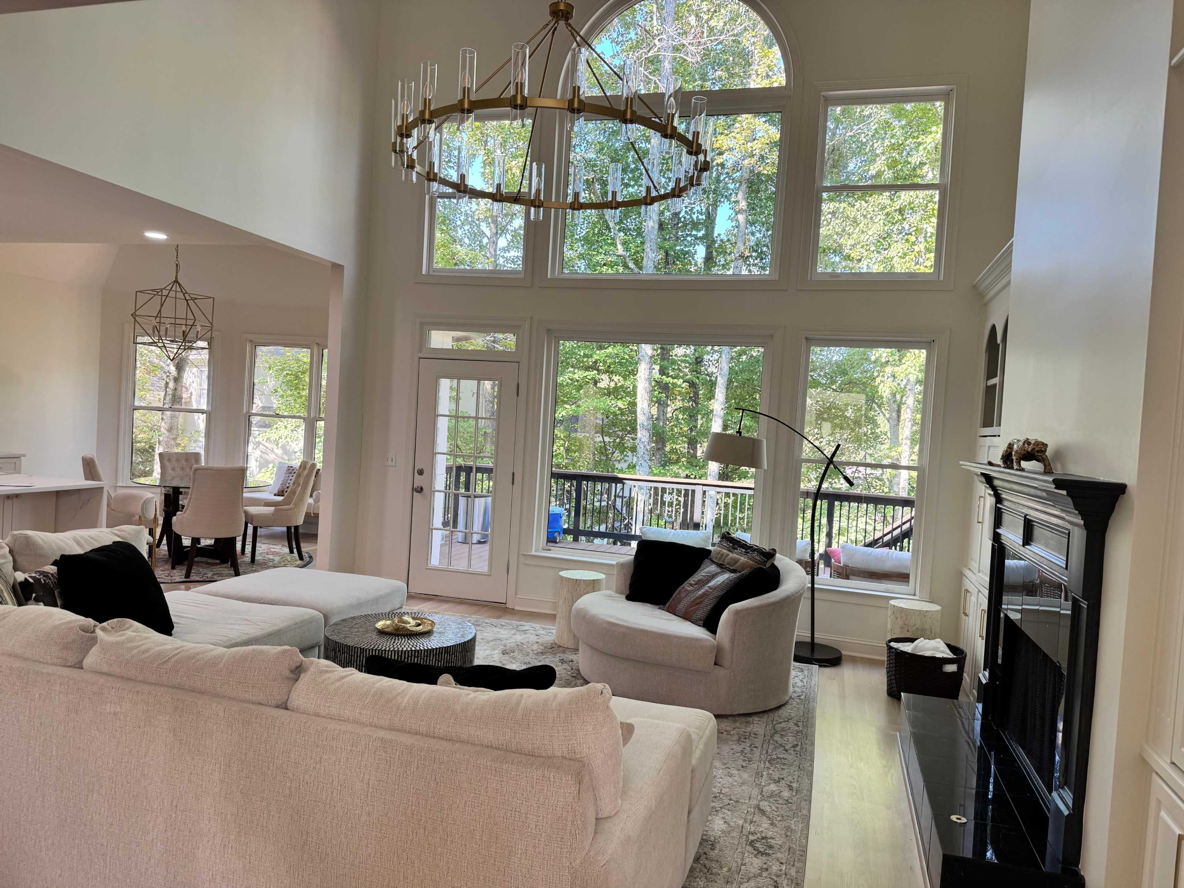 A spacious living room features large windows overlooking a wooded area, with a chandelier hanging above a light-colored sectional sofa and a black fireplace.