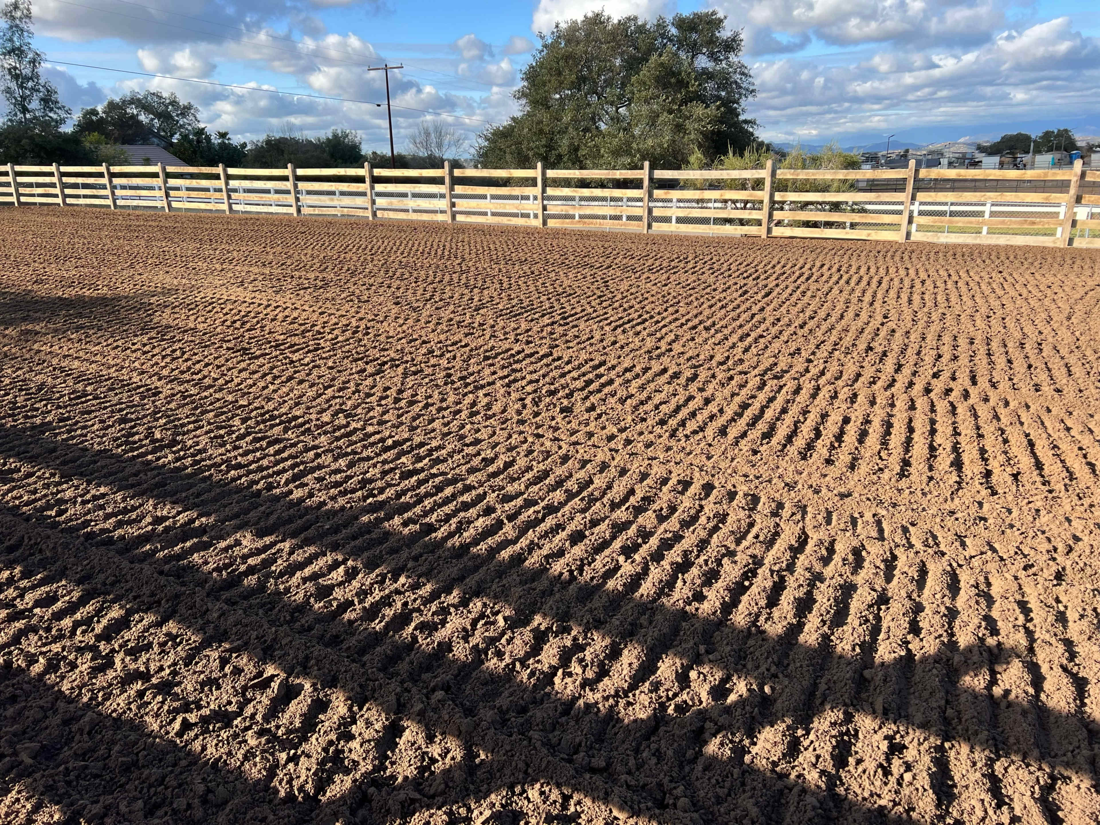 A recently tilled dirt field with clearly defined furrows and a wooden fence in the background.