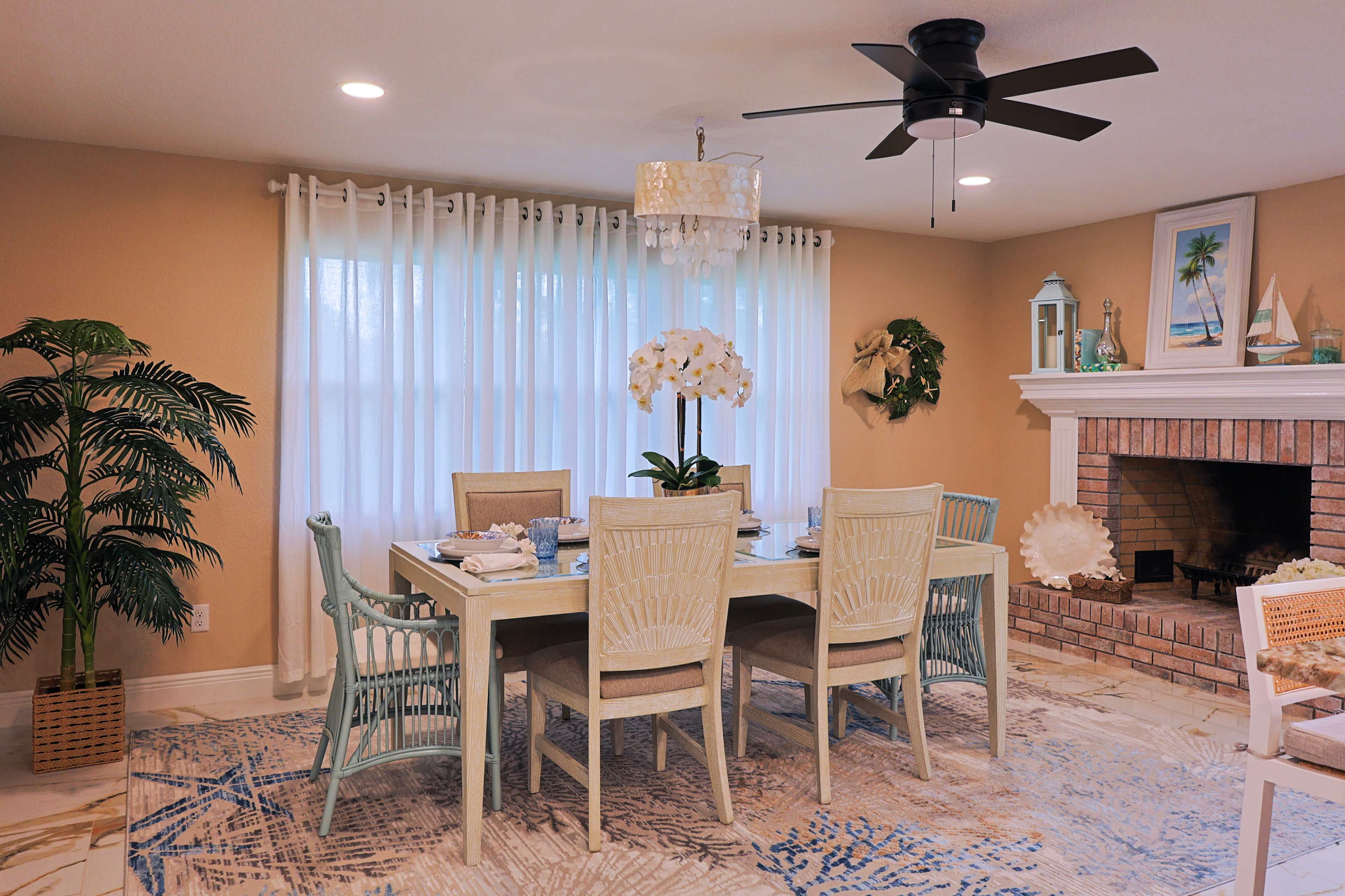 A light and airy dining room features a large table surrounded by six chairs, with a chandelier above and a decorative fireplace on one side.