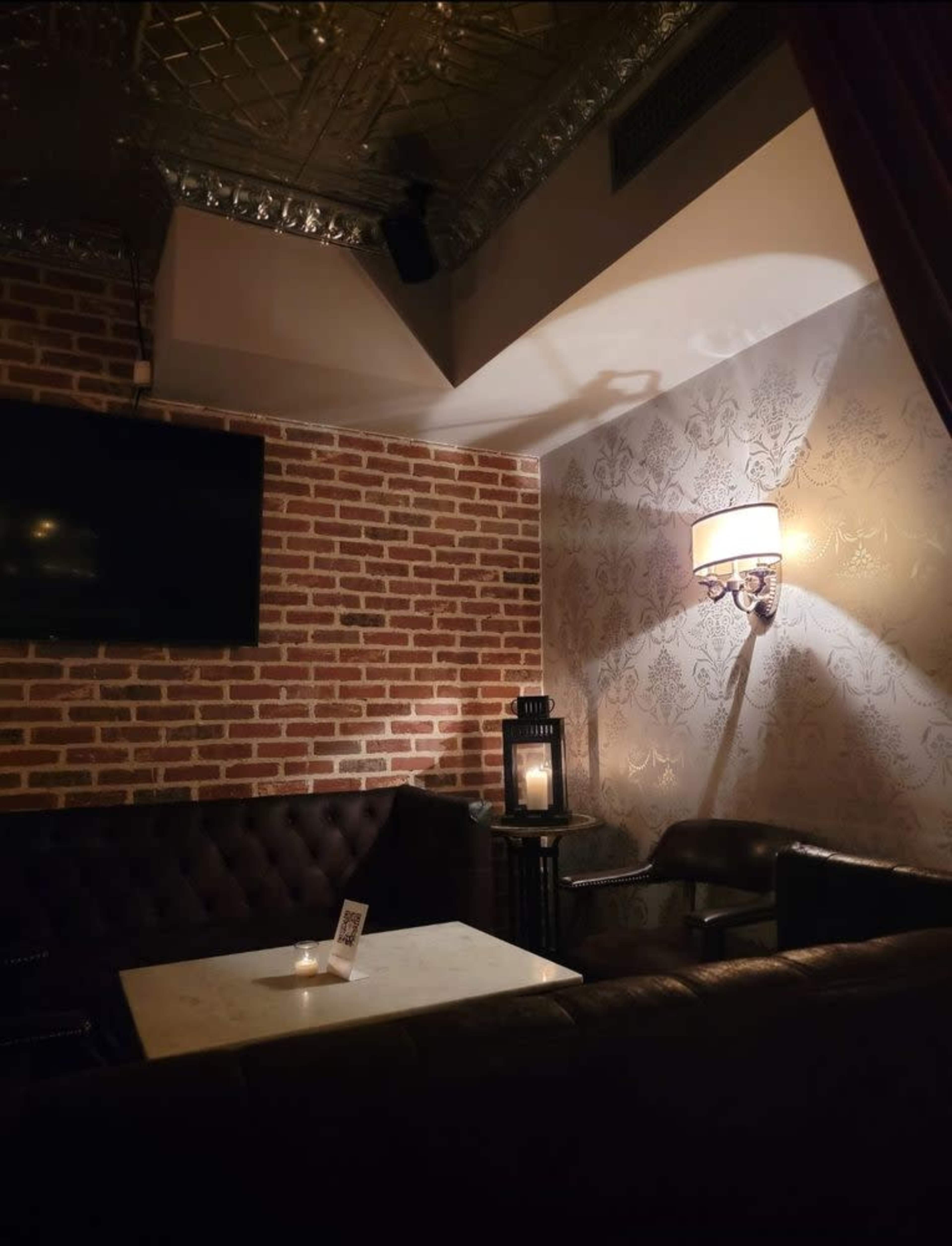 A dimly lit lounge area with a brick wall, a marble table, and a vintage lamp casting light on the patterned wallpaper.