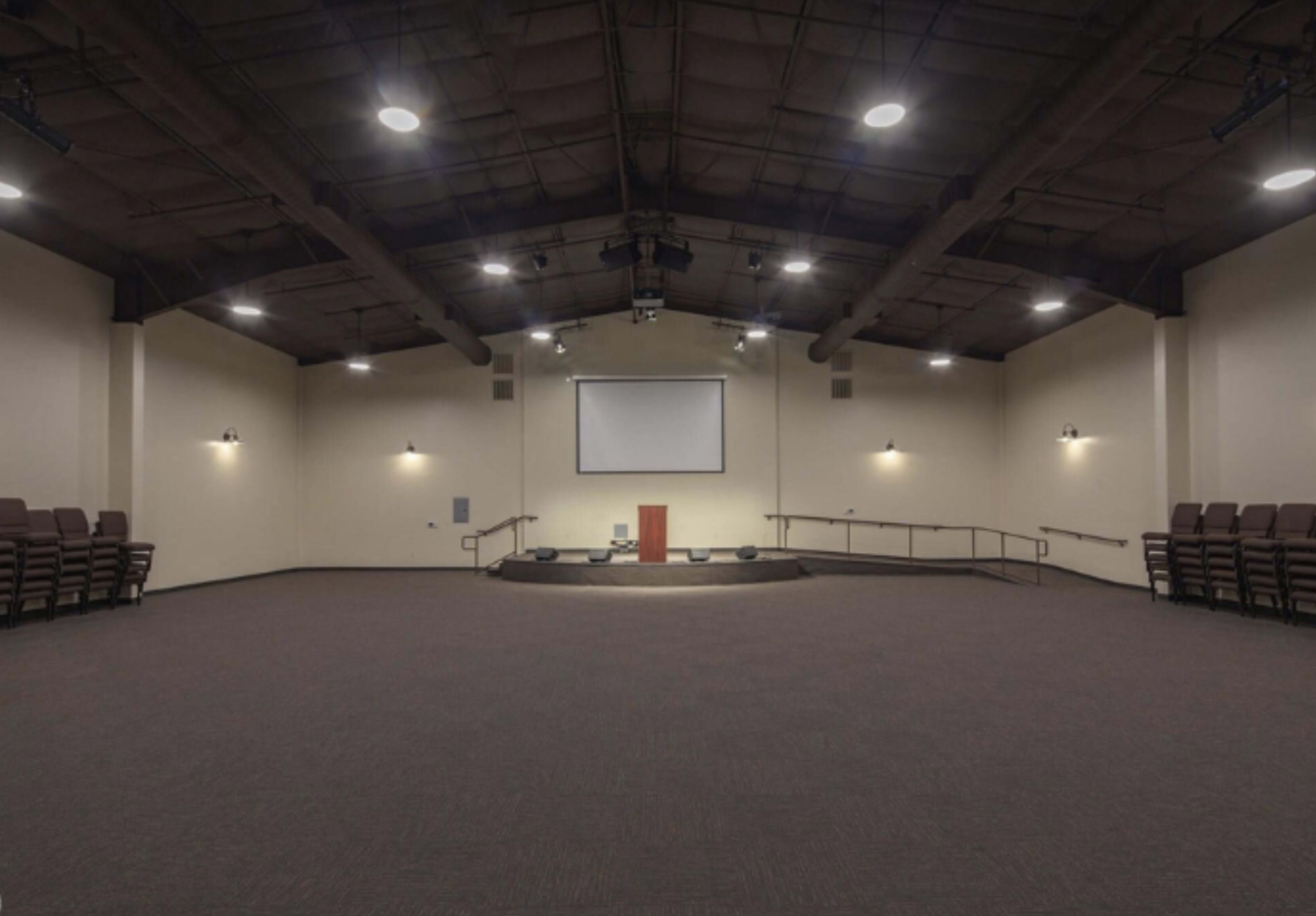 A spacious, empty auditorium with a stage, a screen, and rows of stacked chairs against the walls.
