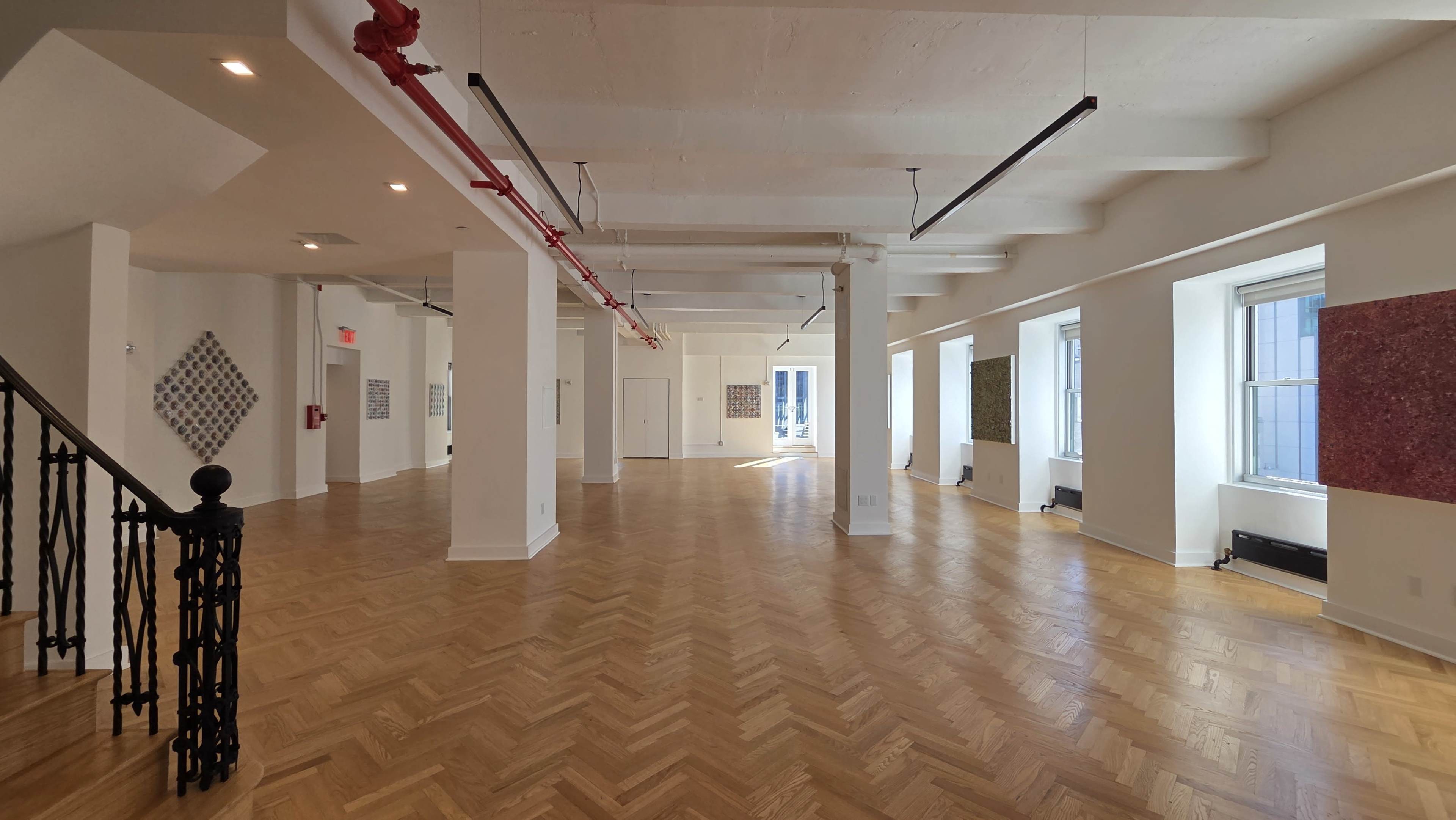 The image shows an empty, spacious room with wooden herringbone flooring, large windows, and white walls, featuring modern lighting and exposed pipes.