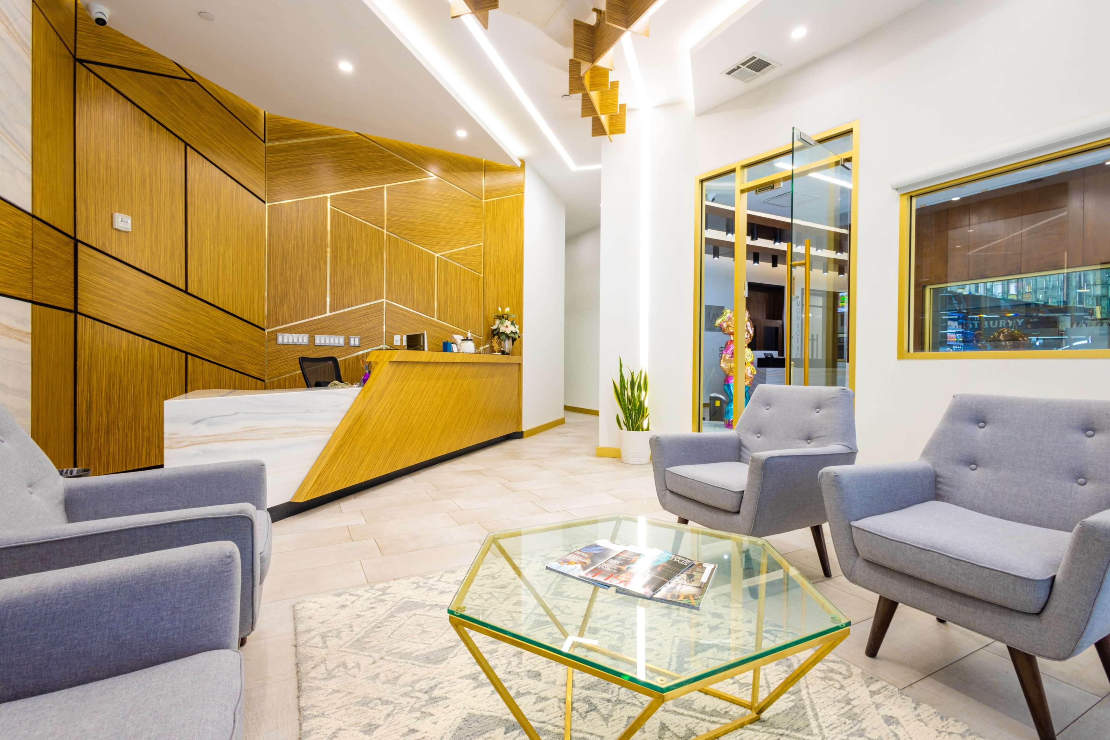 The image shows a modern reception area with wooden accents, grey armchairs, and a glass coffee table on a patterned rug.
