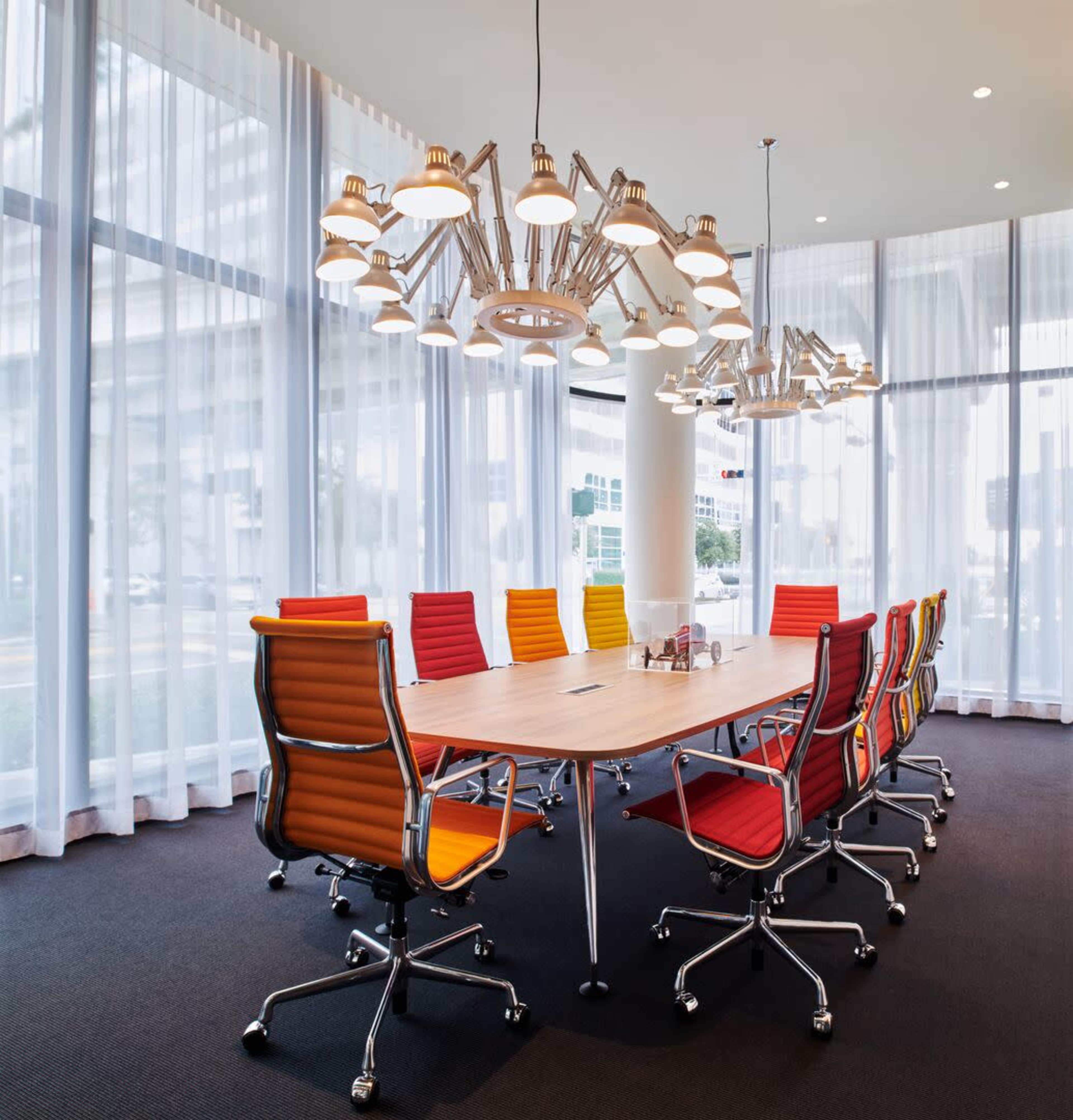 A modern conference room features a large wooden table surrounded by colorful chairs and an overhead light fixture.