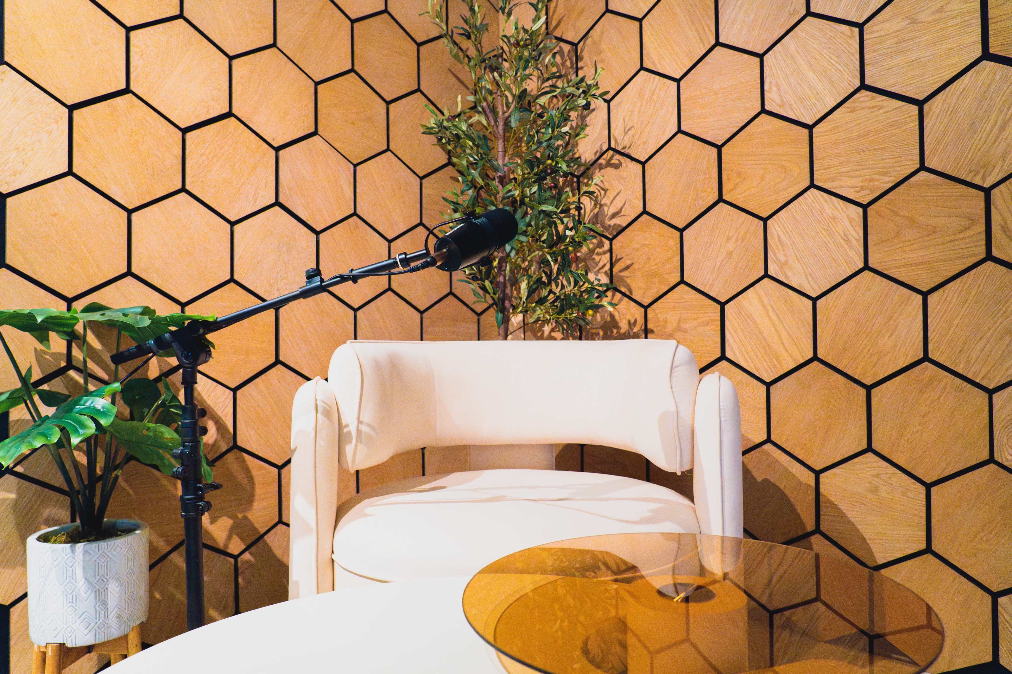 The image shows a modern seating area featuring a white chair, a microphone on a stand, a round glass table, and hexagonal wood paneling on the wall.