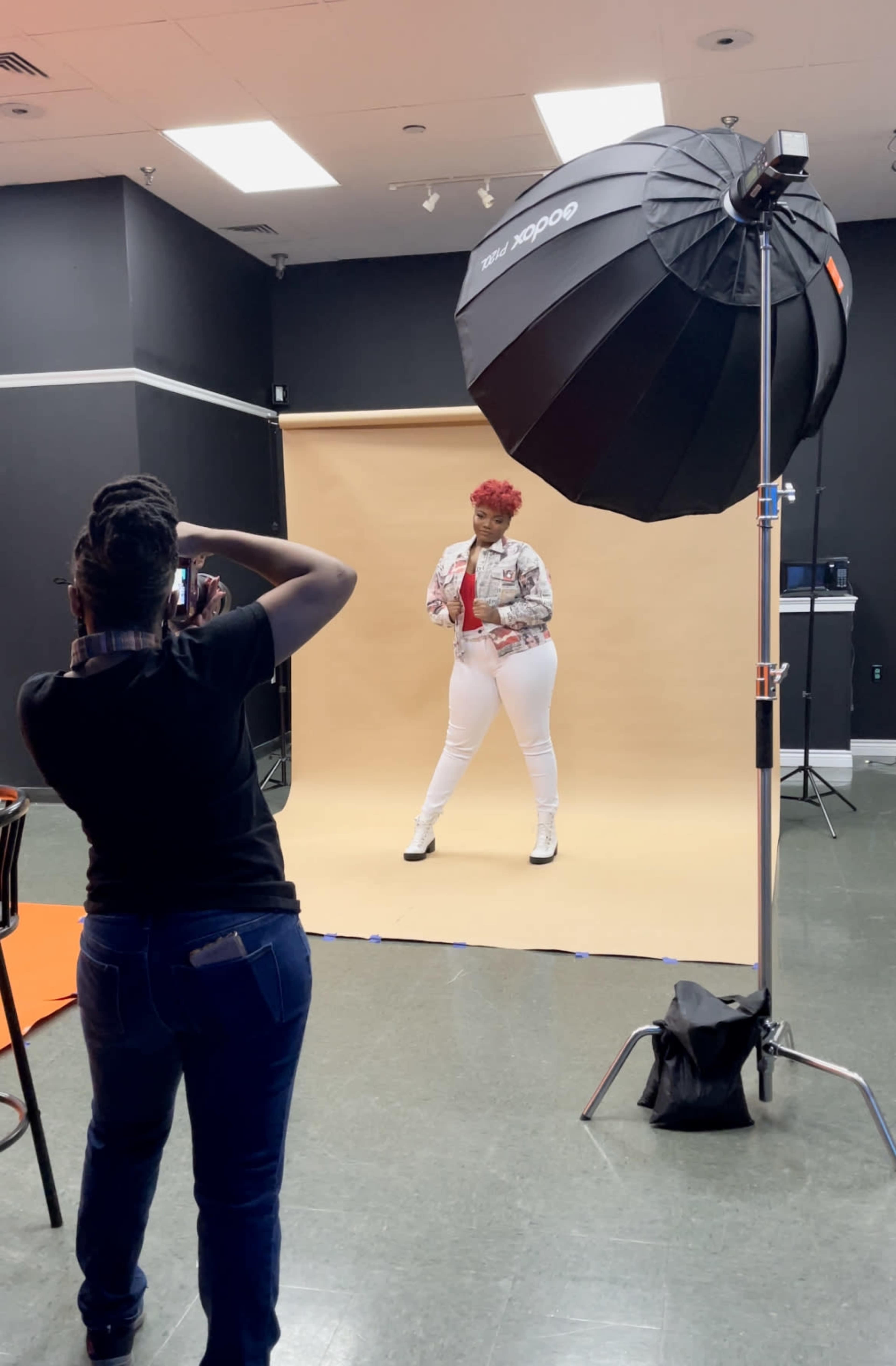 A model poses in front of a beige backdrop while a photographer captures the shot under a studio light.
