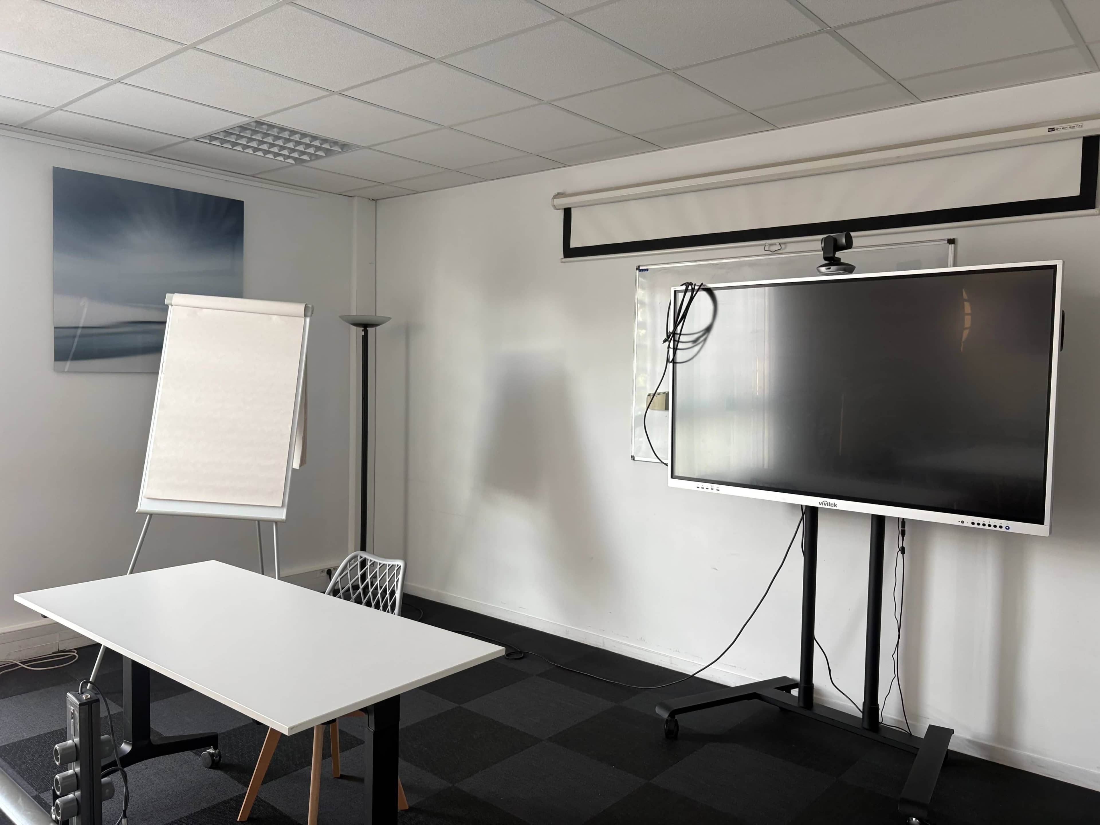 The image shows a meeting room with a large screen, a flip chart, and a white table arranged for a presentation.
