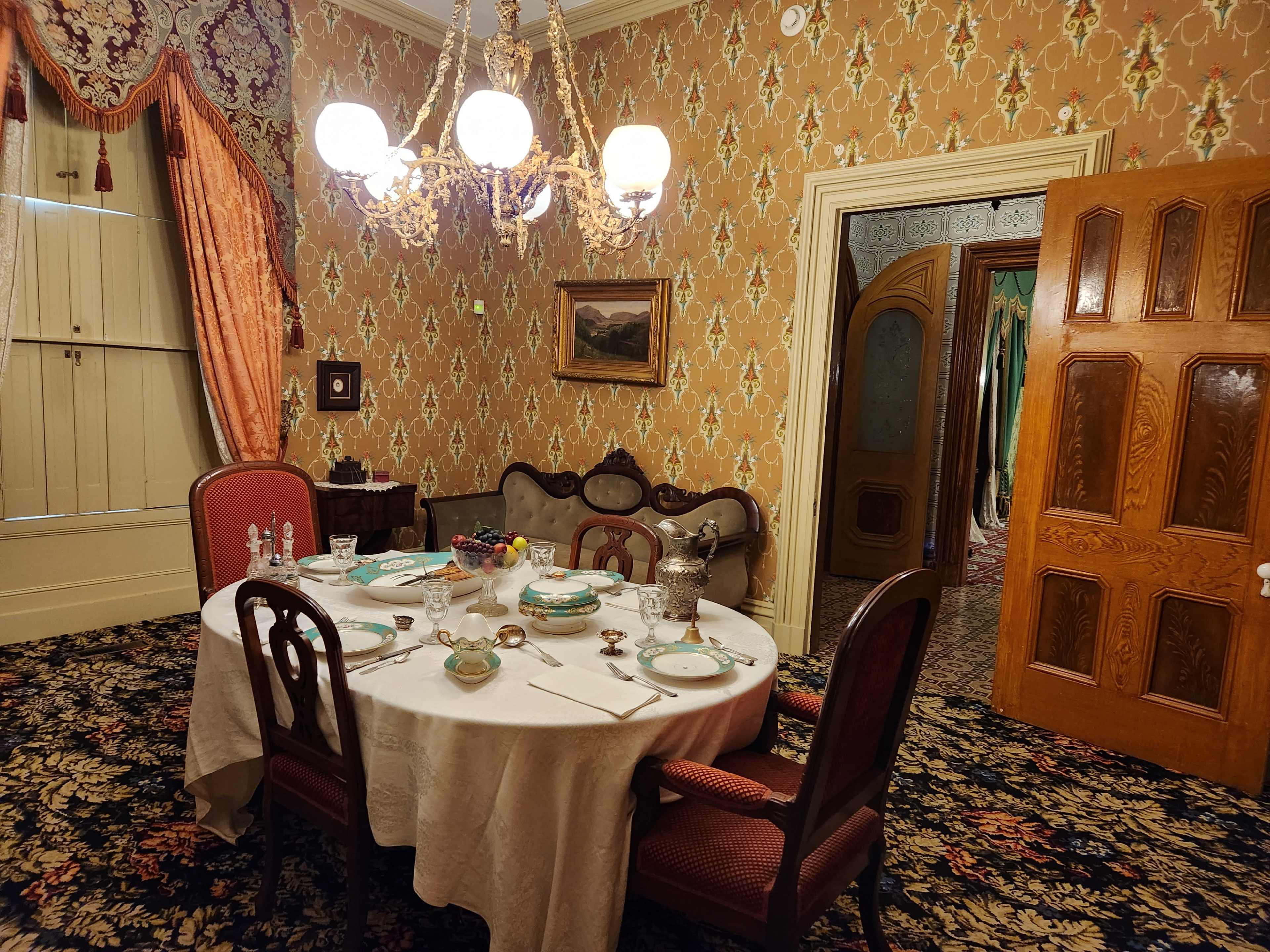 A dining room with a round table set for a meal, featuring vintage decor, patterned wallpaper, and a chandelier.