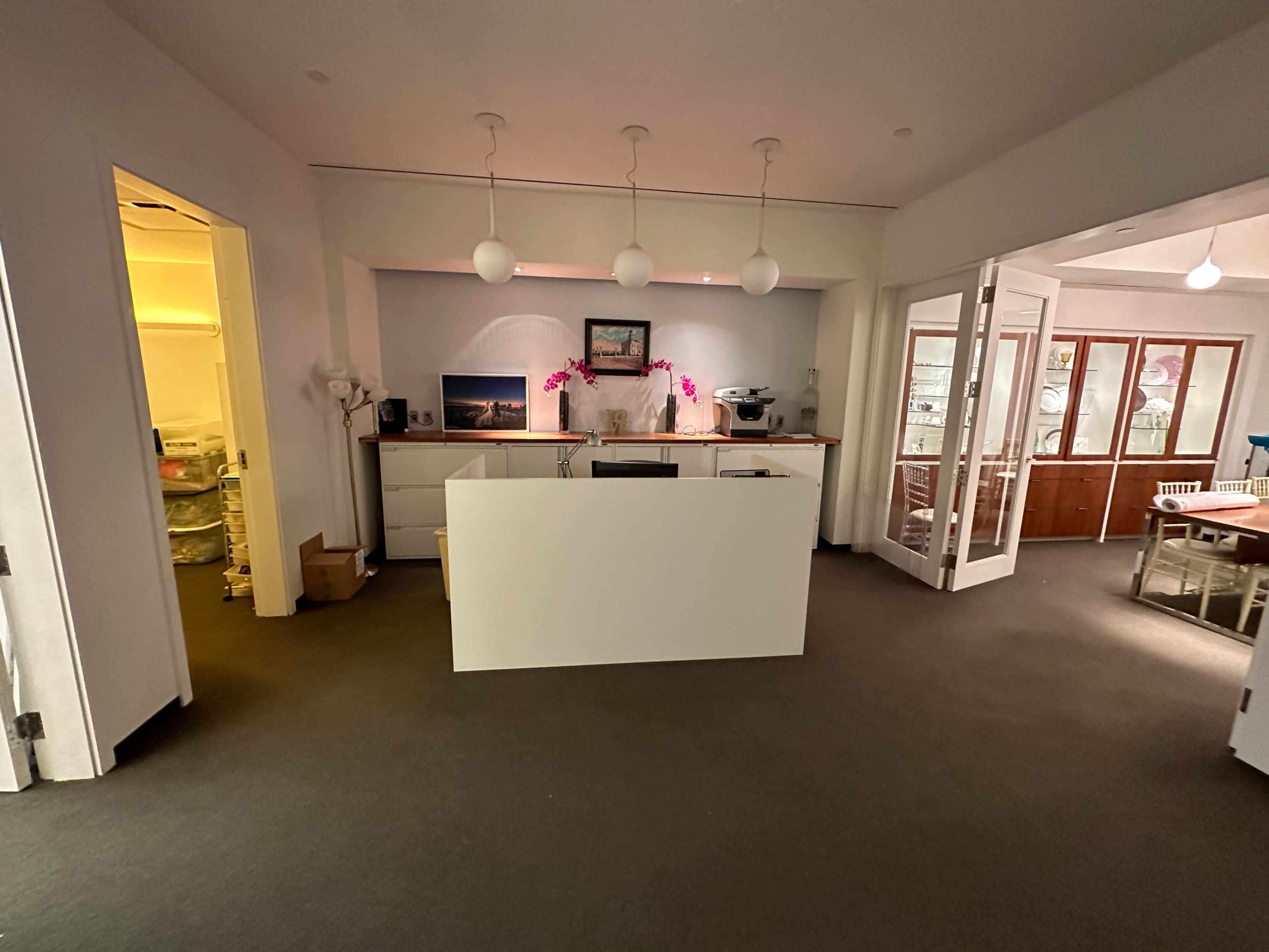 A modern office space with a white reception desk, three hanging lamps, and a view into a room with glass doors and cabinets.