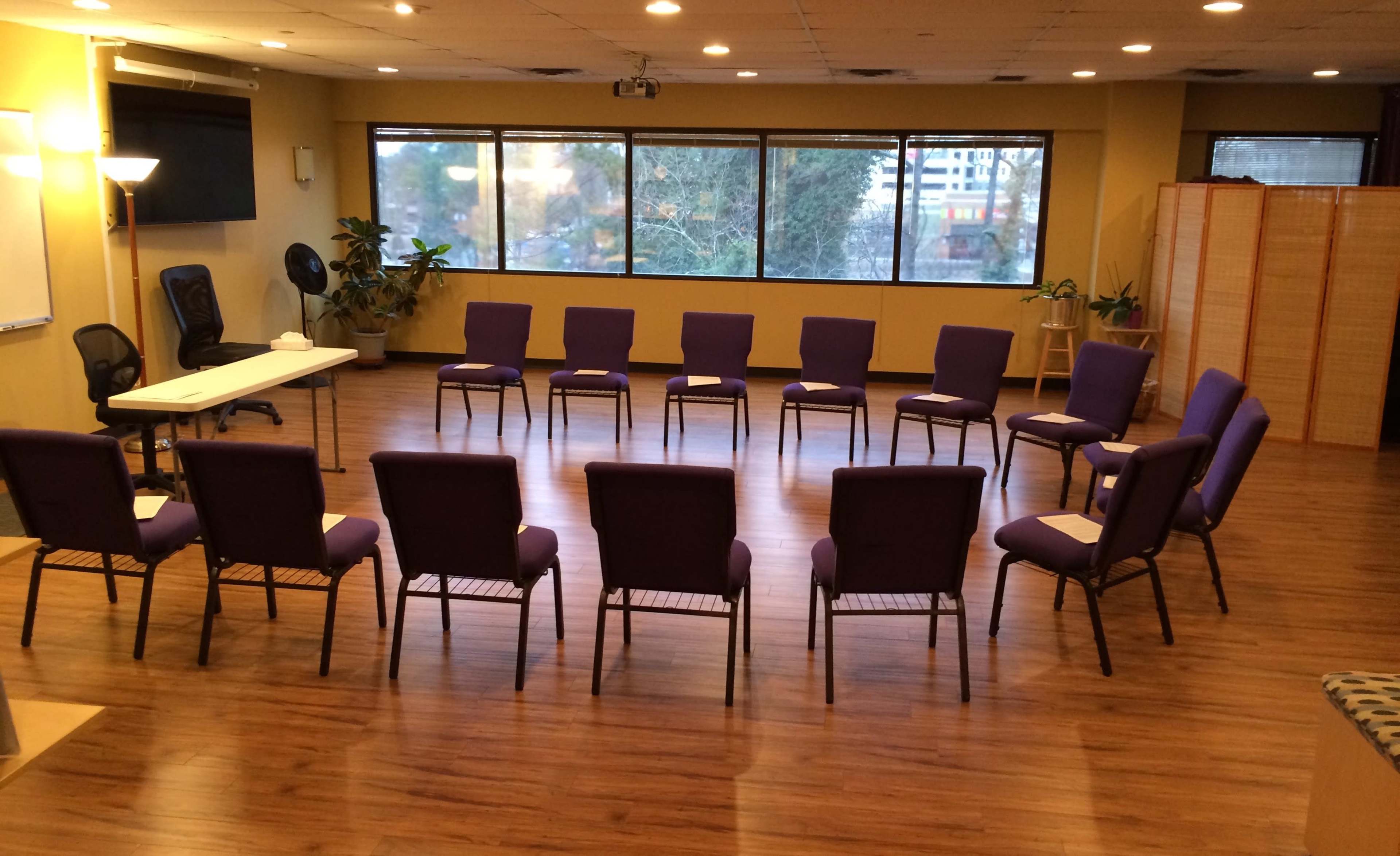 A circular arrangement of purple chairs is set up in a spacious room with large windows and wood flooring.