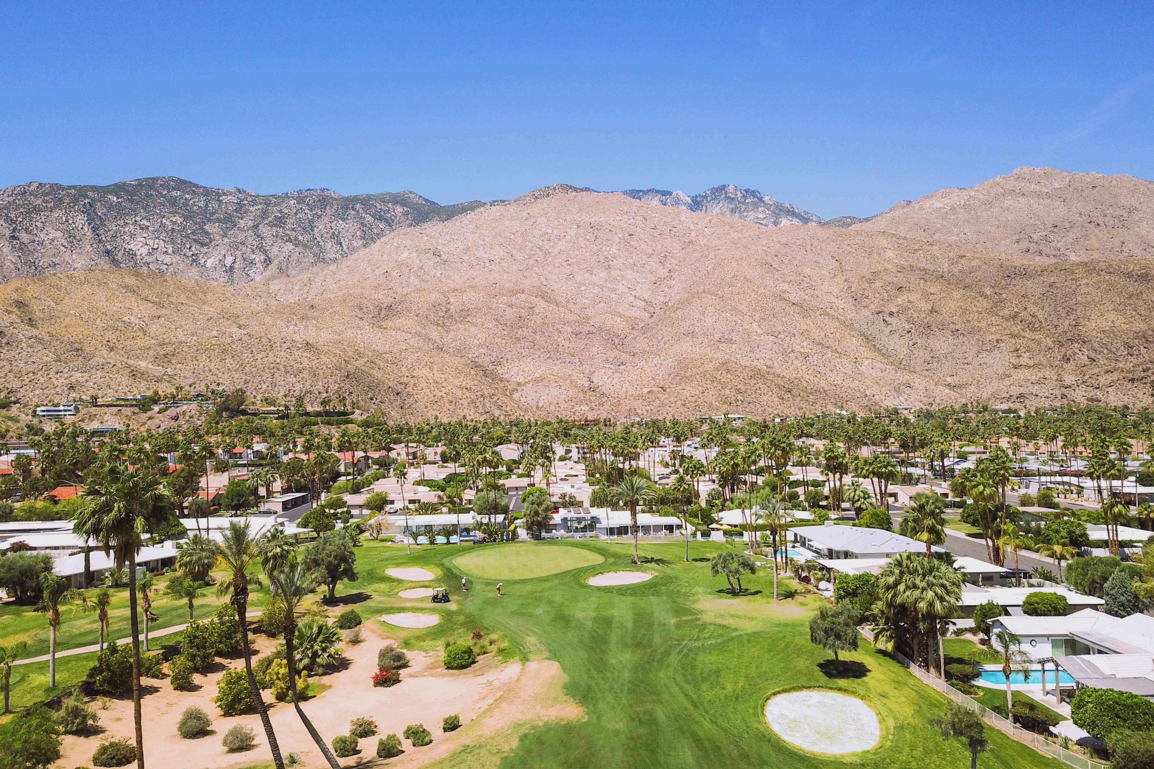 A sprawling golf course surrounded by palm trees and residential buildings, with a mountainous landscape in the background.