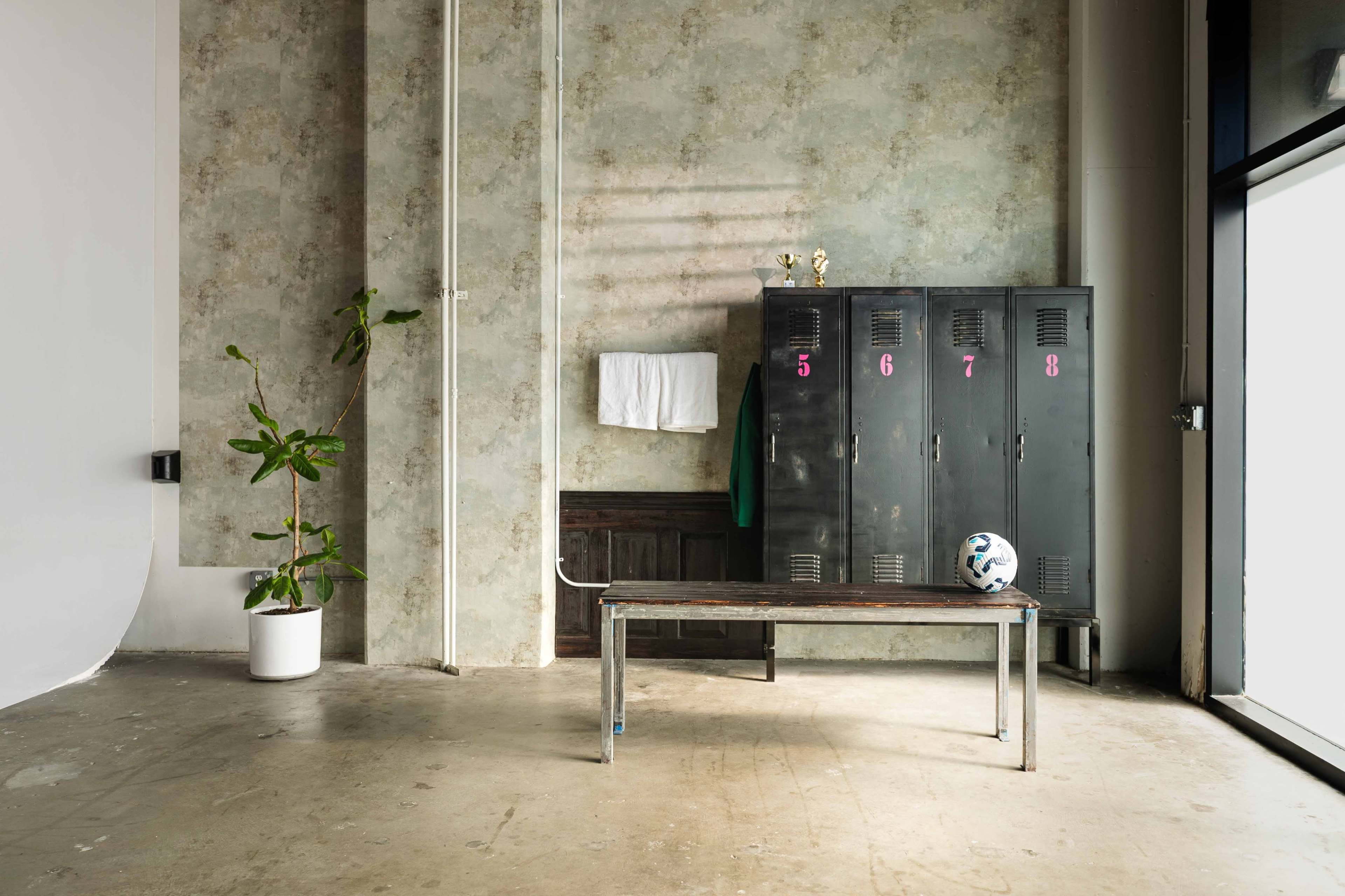 The image shows a locker area featuring a row of black lockers with numbers, a bench, a soccer ball on the floor, and a plant in a white pot against a textured wall.