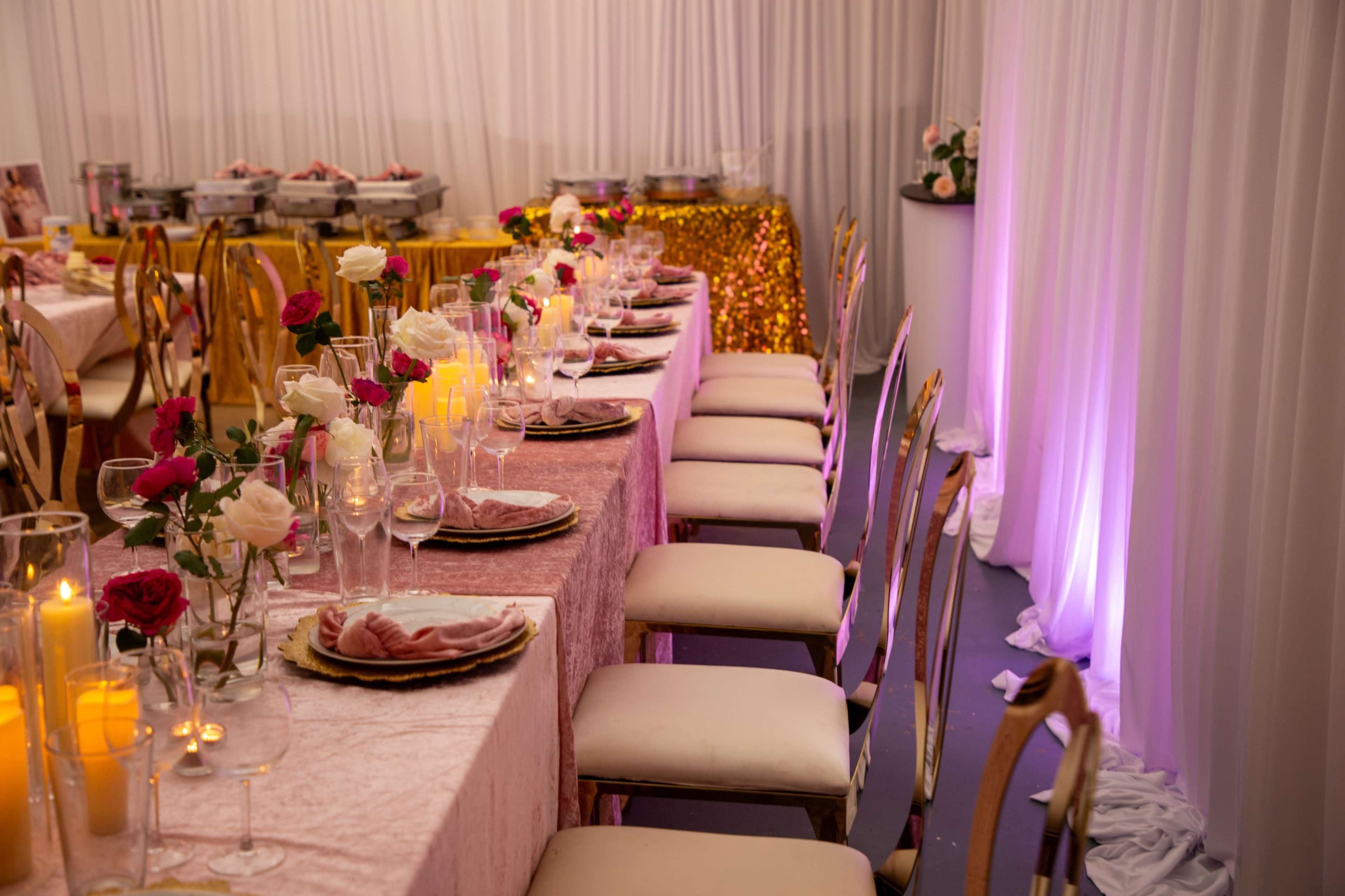 A long dining table is set with decorated plates, glasses, and candles, surrounded by chairs and adorned with flowers in a brightly lit venue.