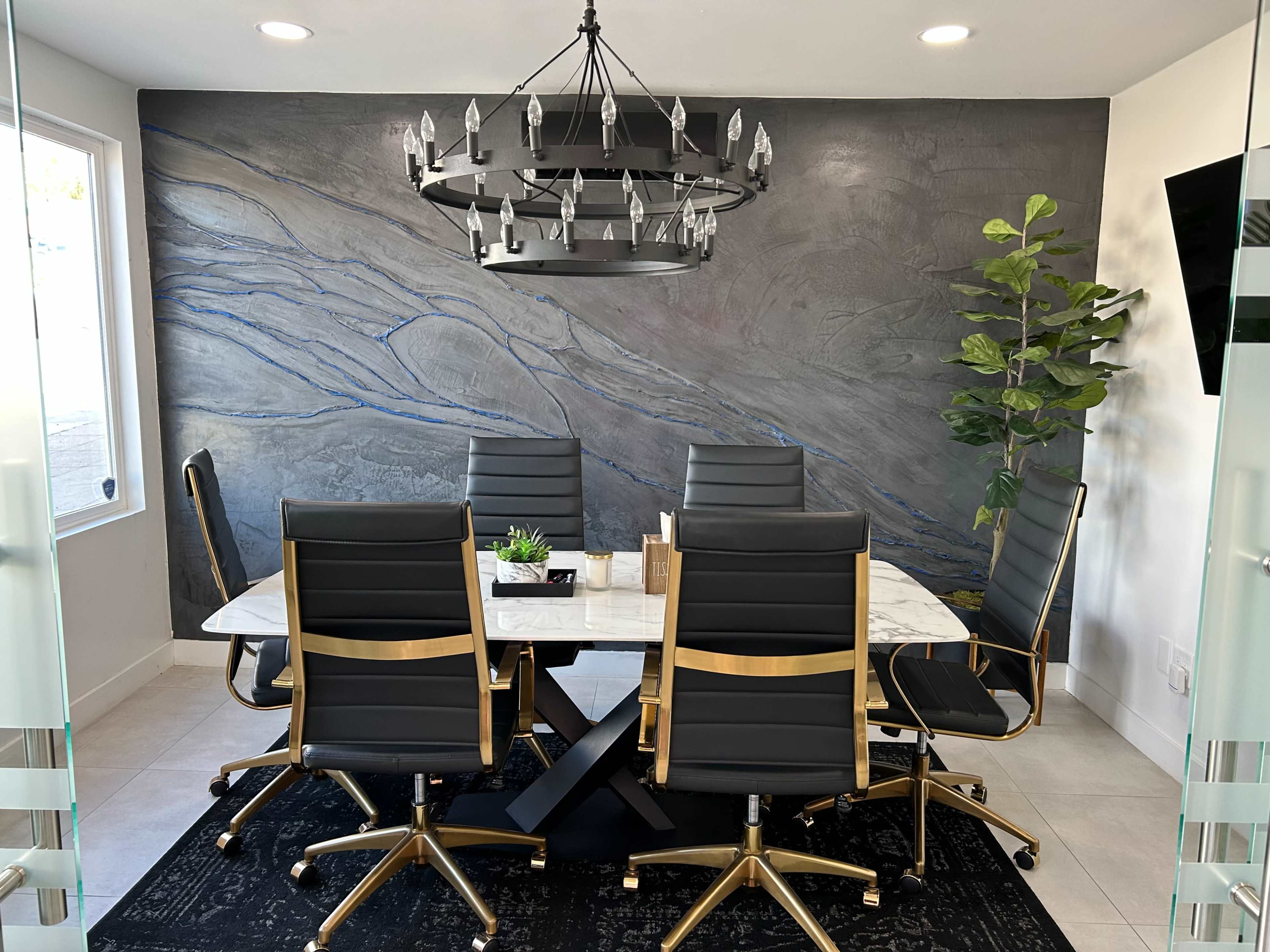 A modern conference room features a large marble table surrounded by sleek black chairs, with a chandelier above and a textured gray wall adorned with blue accents.