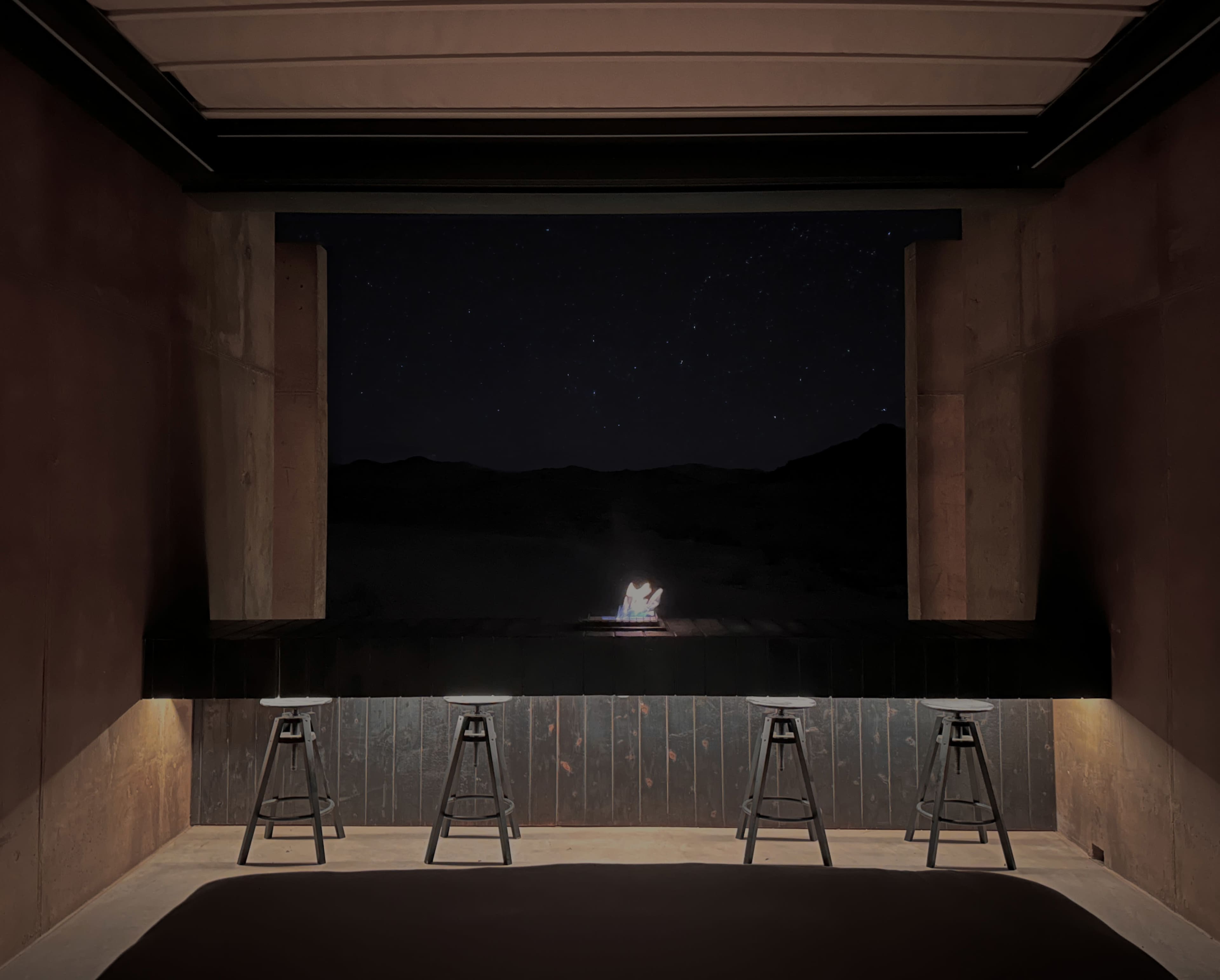 A dimly lit interior space with three stools facing an open window, which frames a distant campfire under a starry night sky.