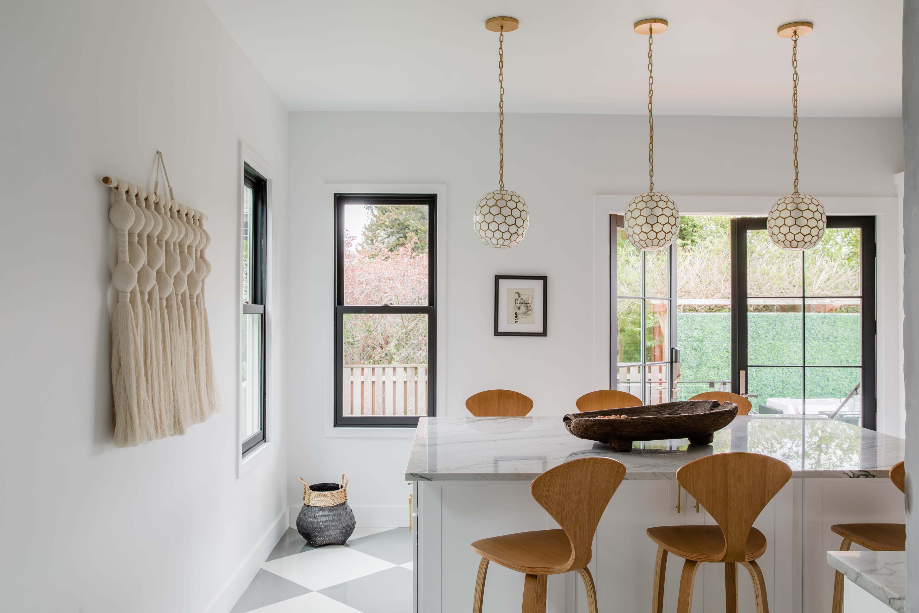 A modern kitchen features a marble island with wooden stools, pendant lights, and large windows overlooking an outdoor area.