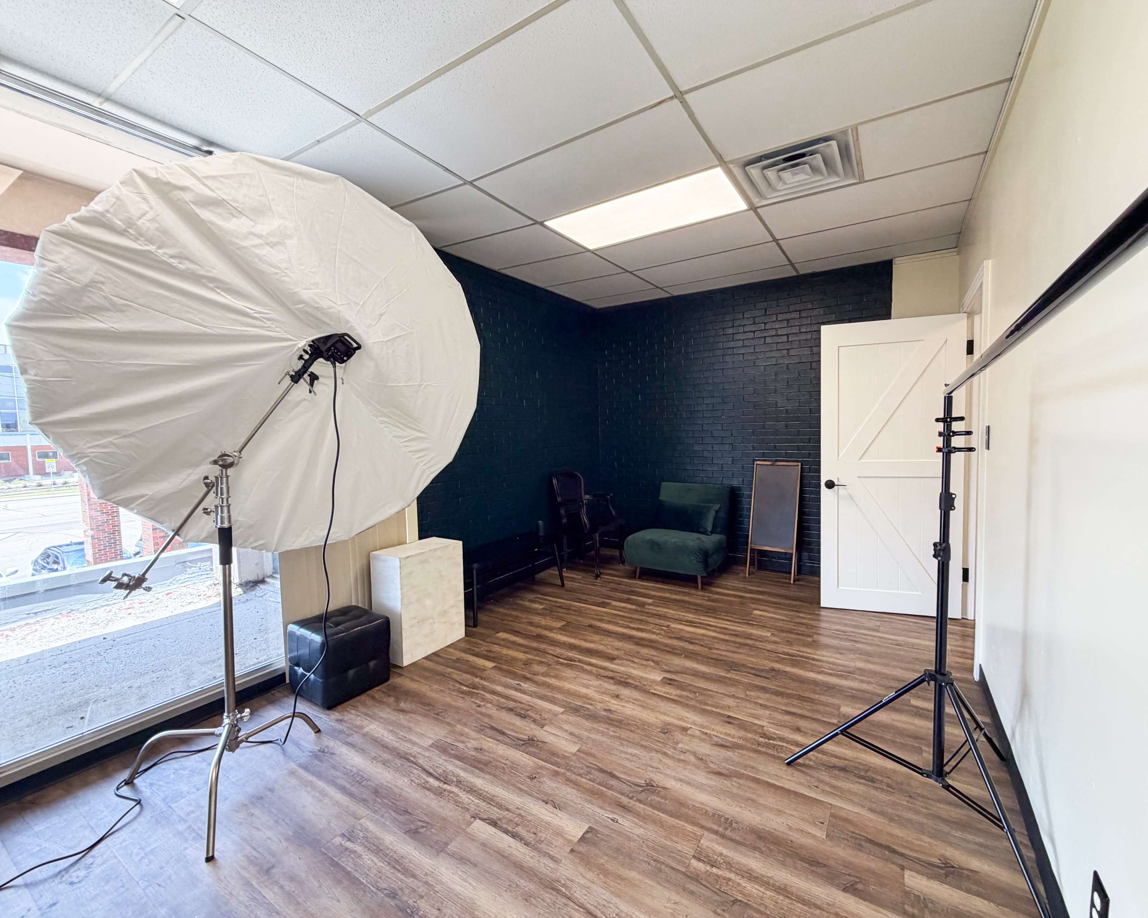 The image shows a spacious, well-lit room featuring a large softbox light, a green couch, a chair, and a mirror against a dark wall, with a door and white backdrop stand visible.