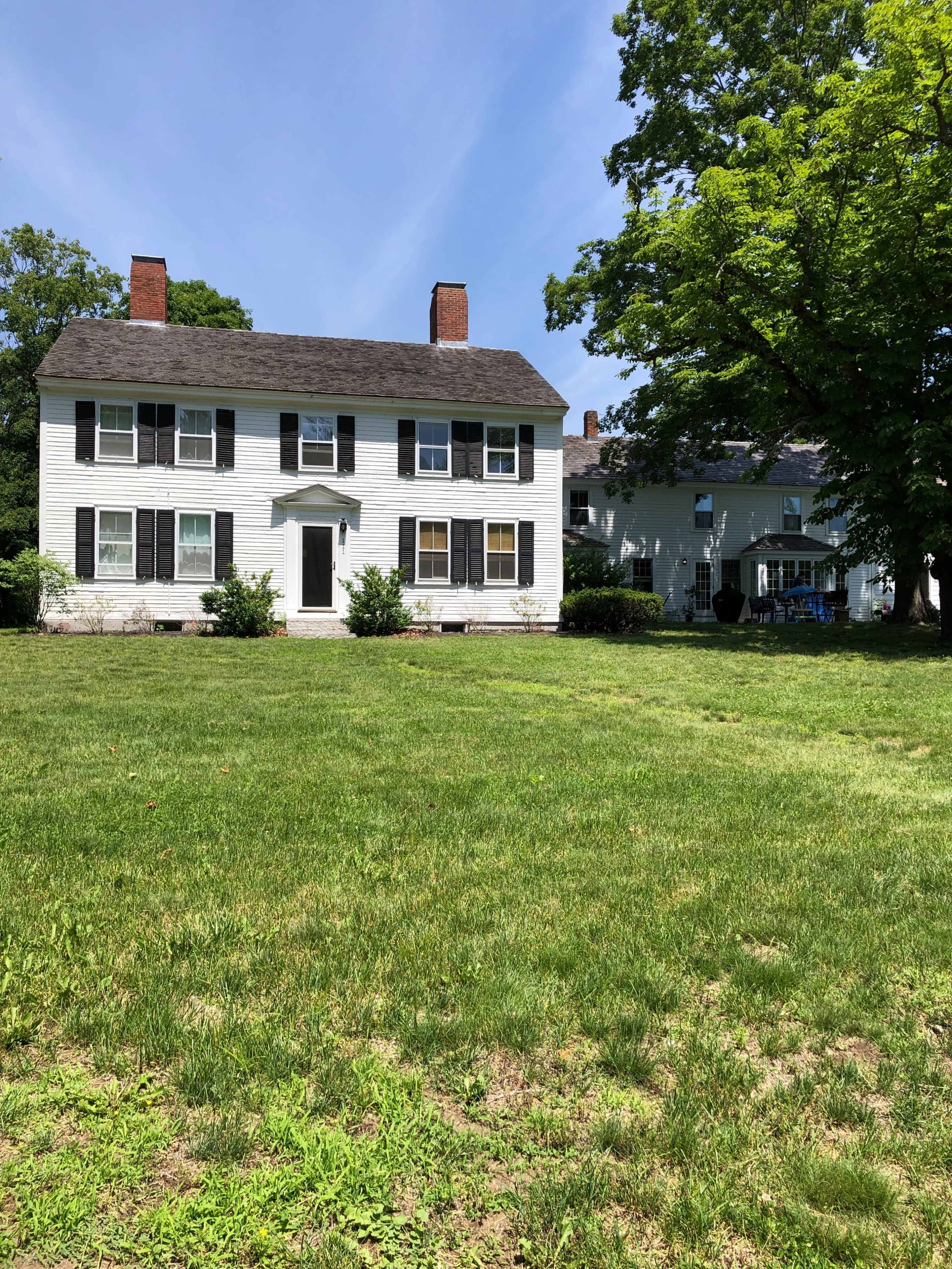 A two-story white colonial house with black shutters, situated on a grassy lawn, surrounded by trees.