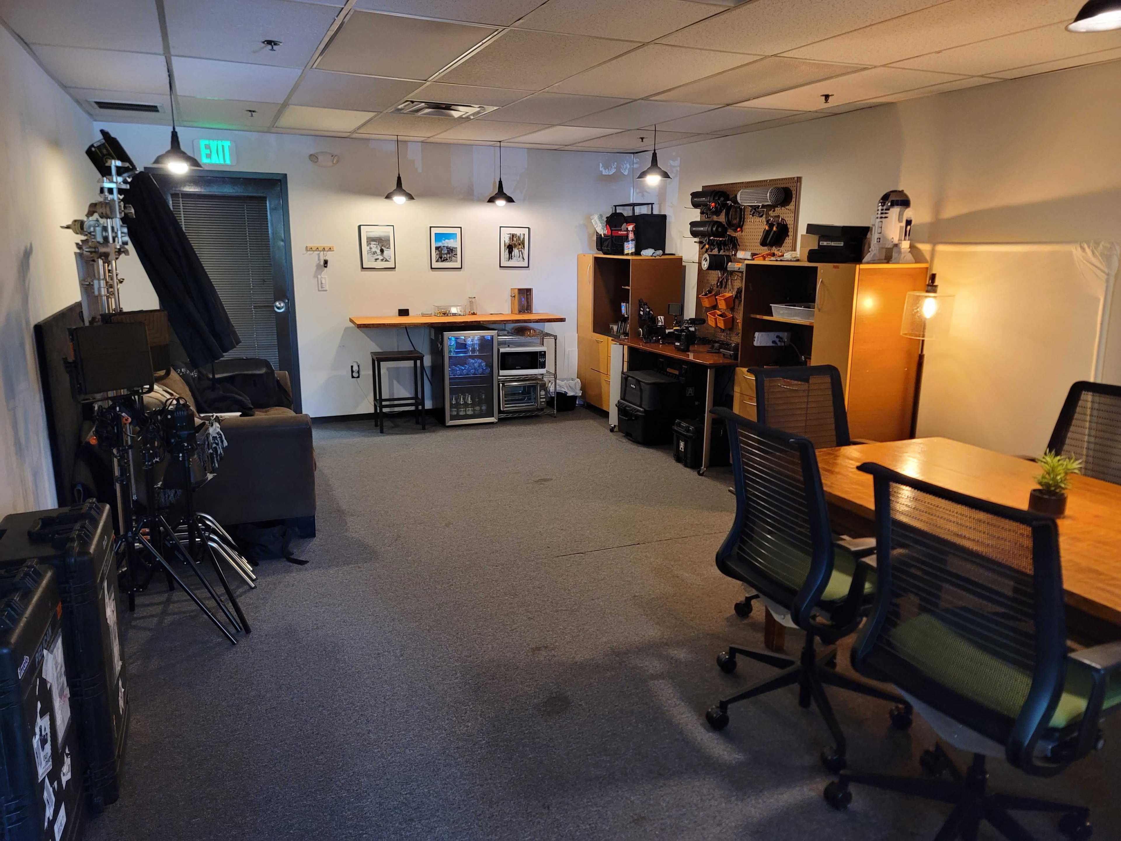 An office space with a meeting table, several technology setups, storage cabinets, and a mini refrigerator.