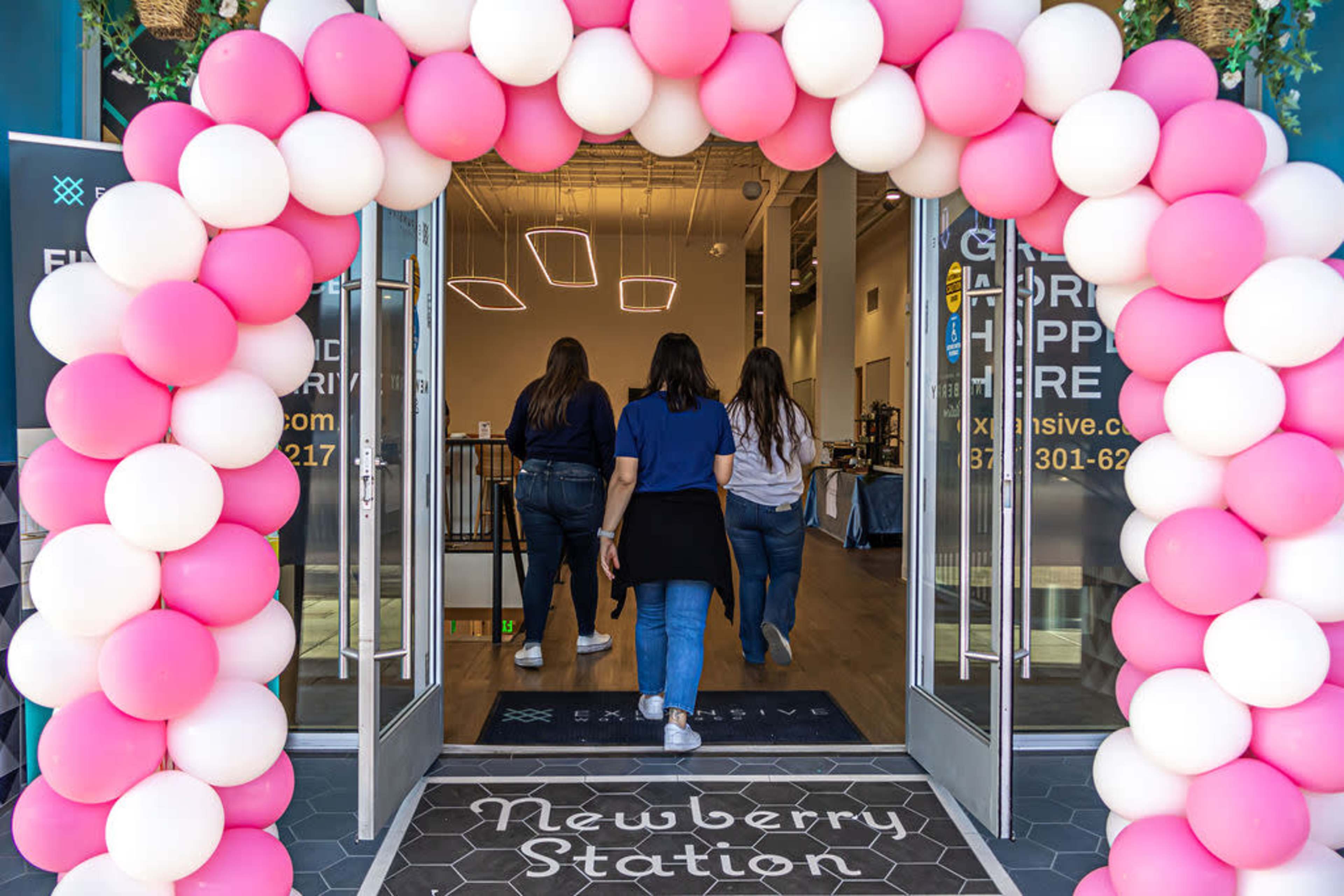 A group of four people walks through a colorful balloon archway into a building labeled "Newberry Station."