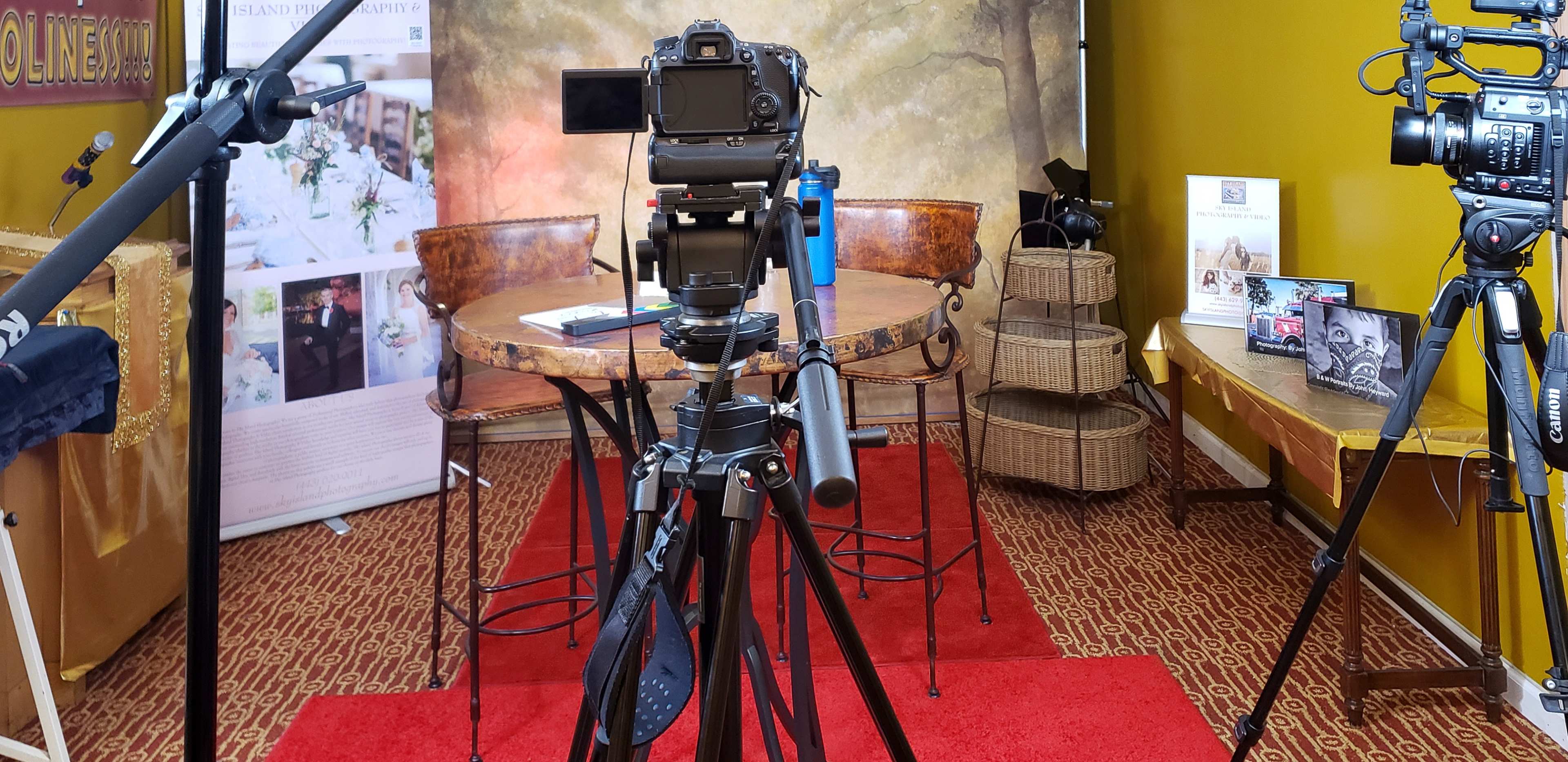A setup for a video recording session with multiple cameras, tripods, and a decorated background featuring a table and chairs.