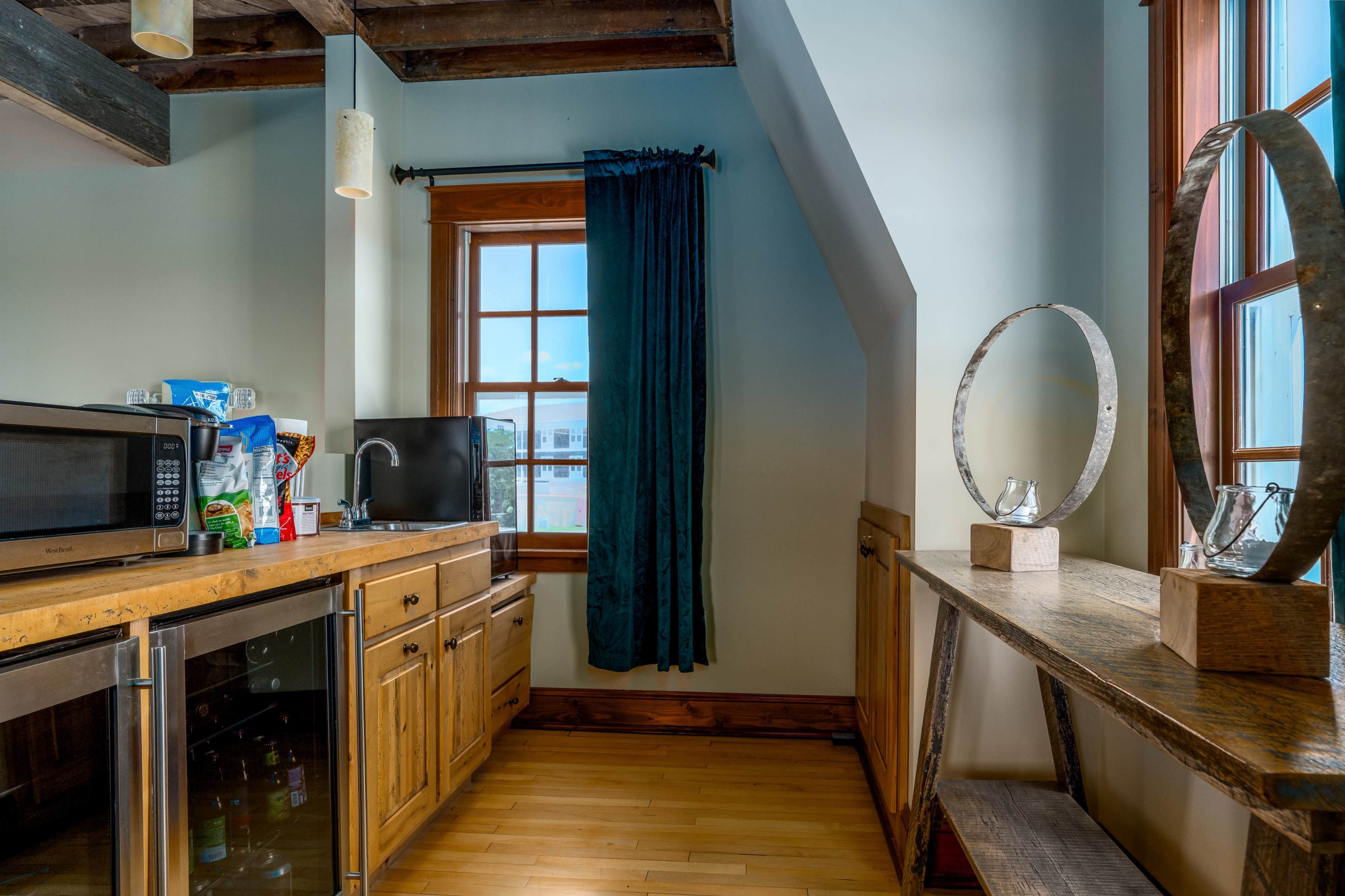 A kitchen corner with a microwave, sink, wooden cabinets, and a shelf displaying decorative items, all under a window with blue curtains.