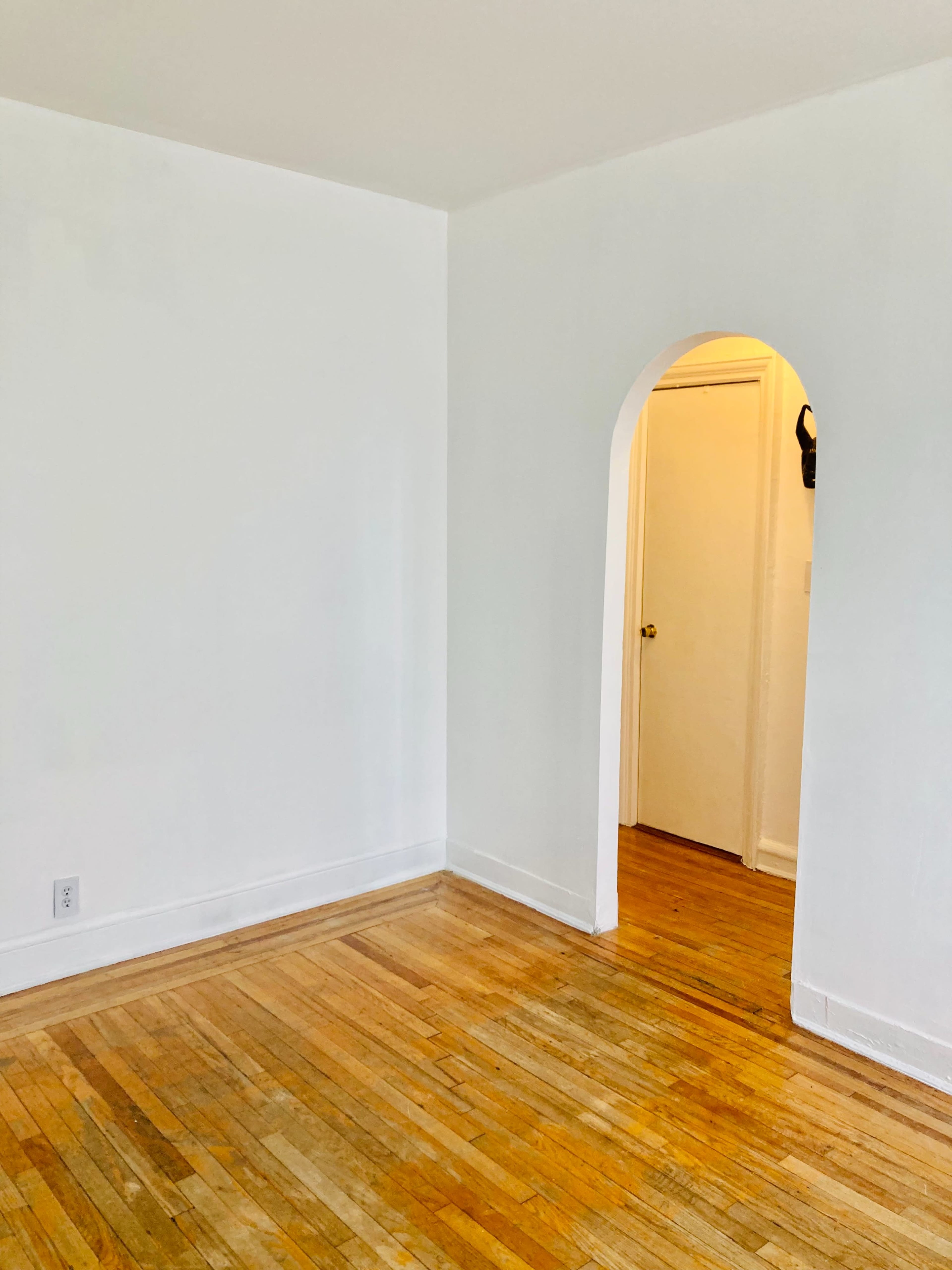 A partially empty room with light-colored walls and wooden flooring, featuring an arched doorway leading to another room.