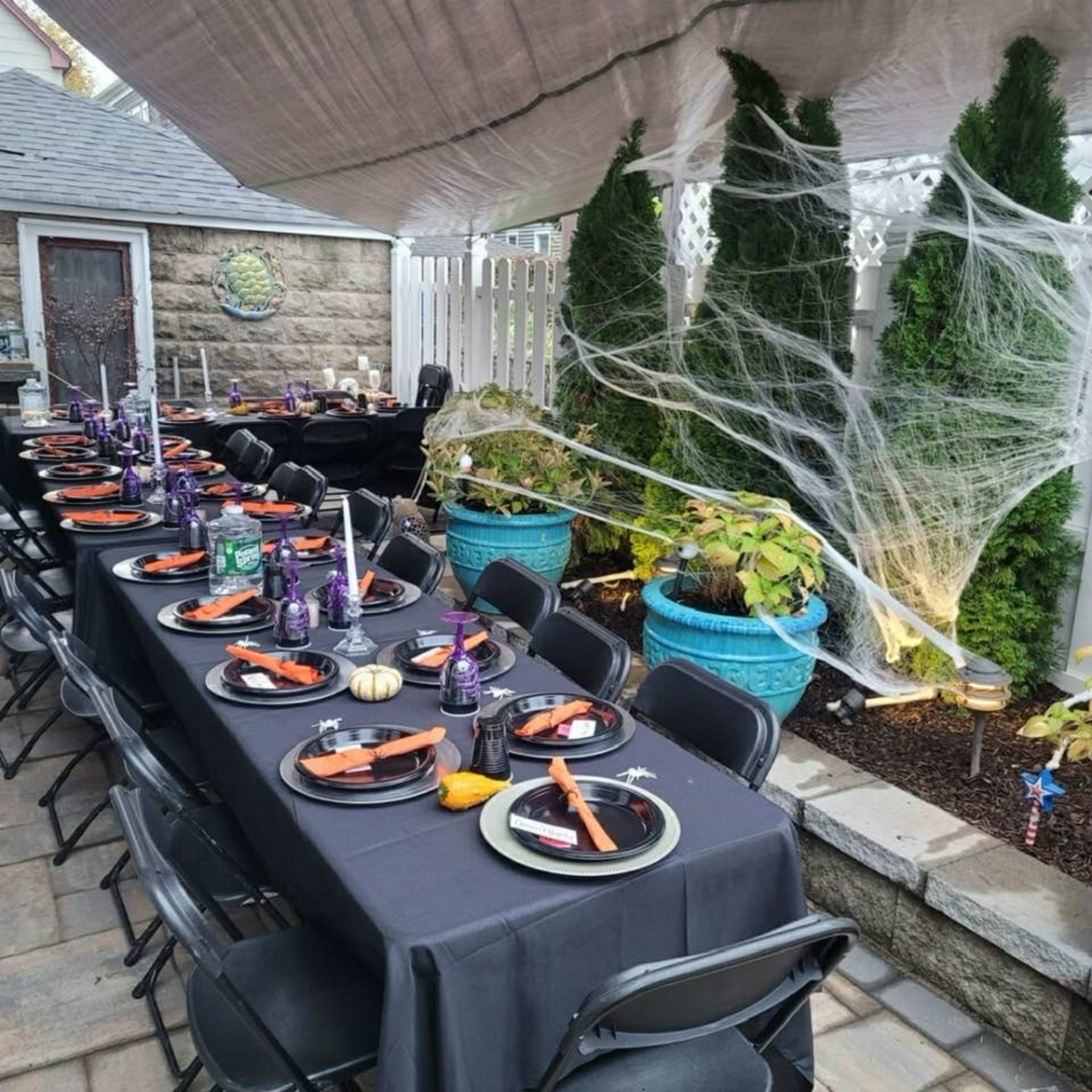 The image shows a long outdoor dining table set with black tablecloths and Halloween-themed decorations, including purple and orange accents, surrounded by a garden filled with cobwebs and potted plants.