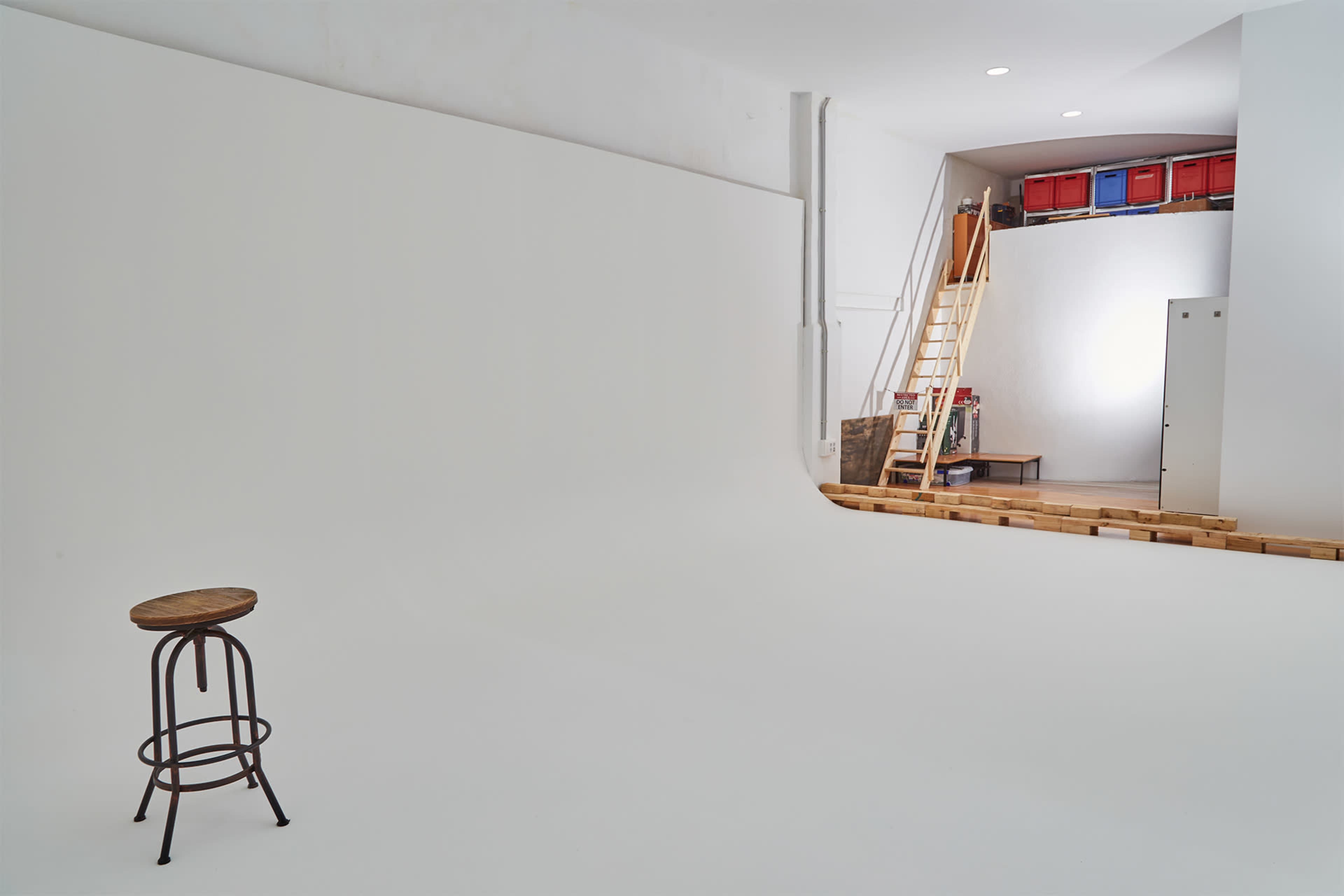 A wooden stool stands alone in a minimalist studio space with a white backdrop and a ladder leading to a storage area.