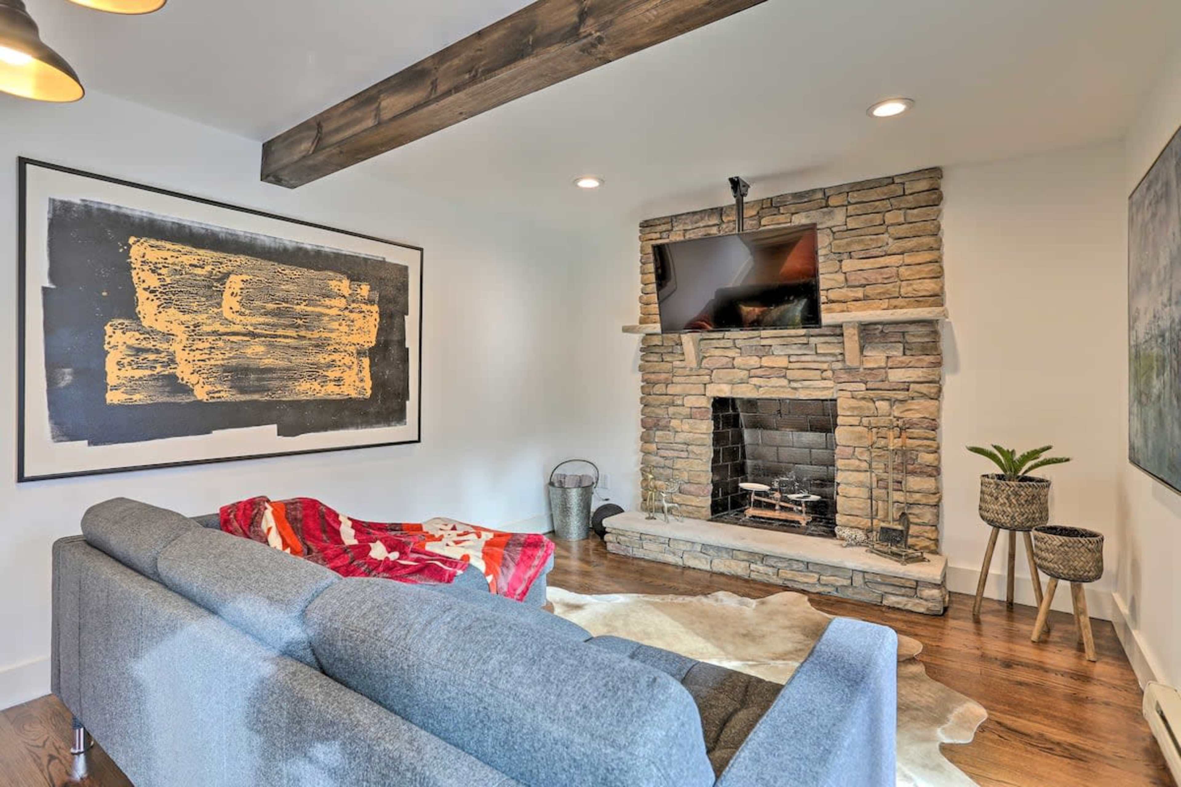A cozy living room features a stone fireplace, a mounted television, a gray sofa with a red throw blanket, and a large framed artwork on the wall.