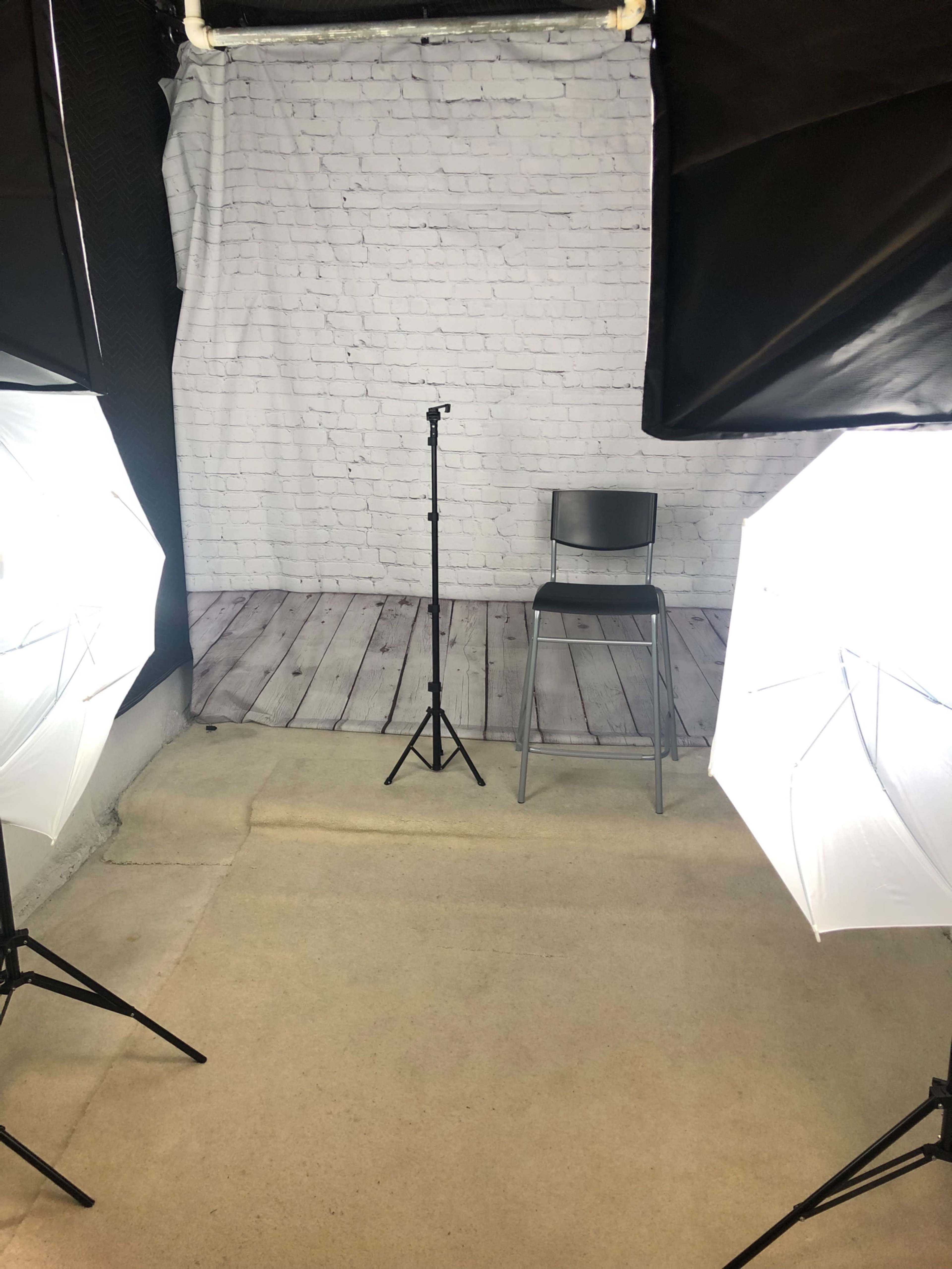 A simple studio setup with a microphone stand and a chair positioned in front of a brick-patterned backdrop, illuminated by two softbox lights.