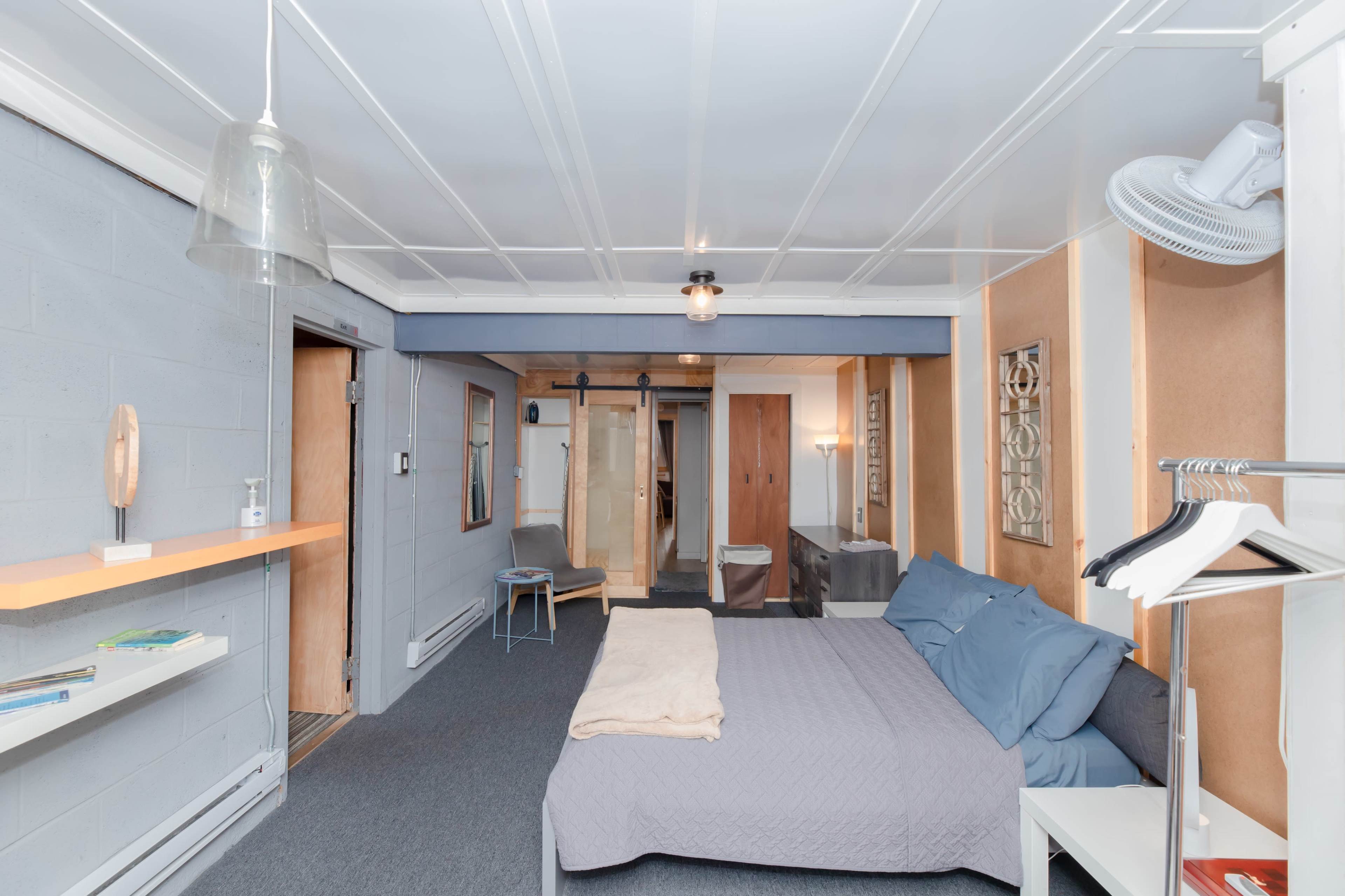 The image shows a compact bedroom featuring a bed with blue bedding, a small seating area, and a combination of wooden and gray walls.