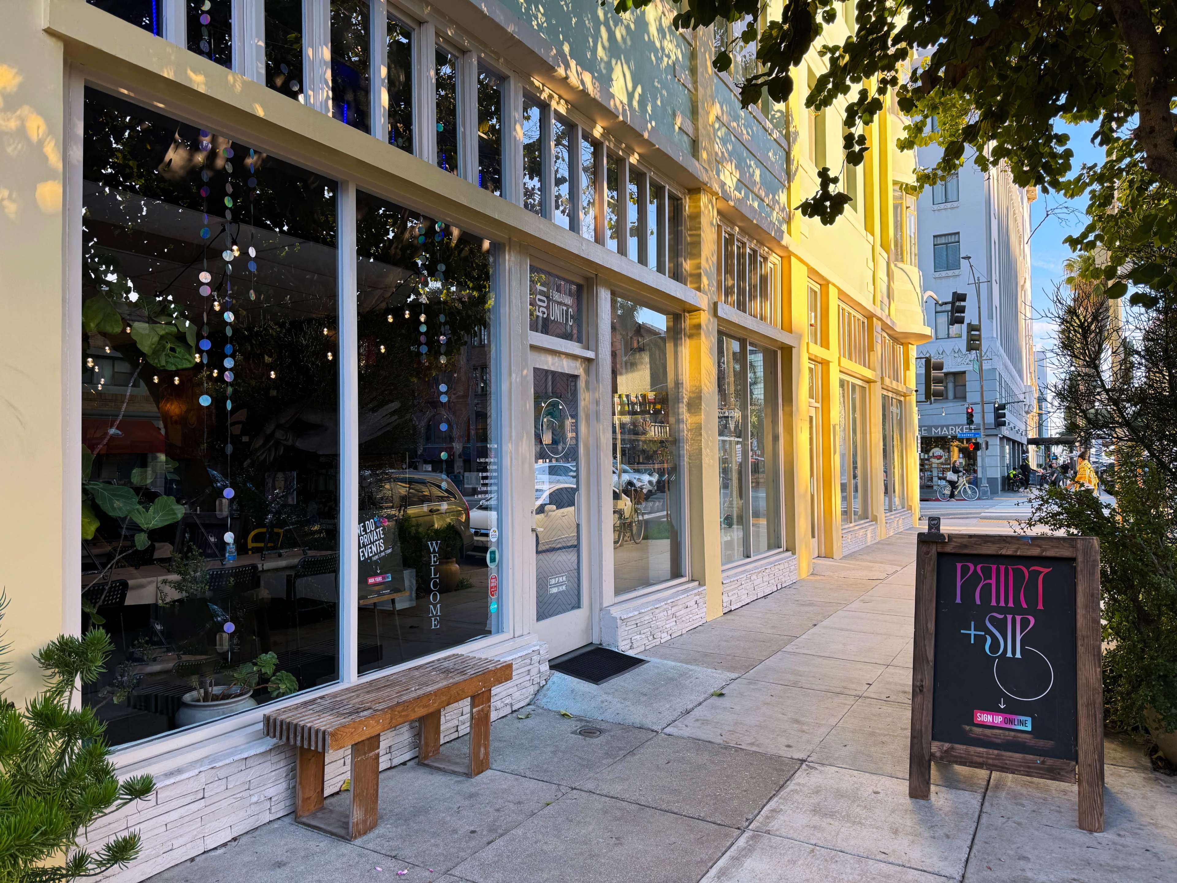 A storefront of a business named "Paint + Sip," with large windows displaying art supplies and a sidewalk lined with a wooden bench.