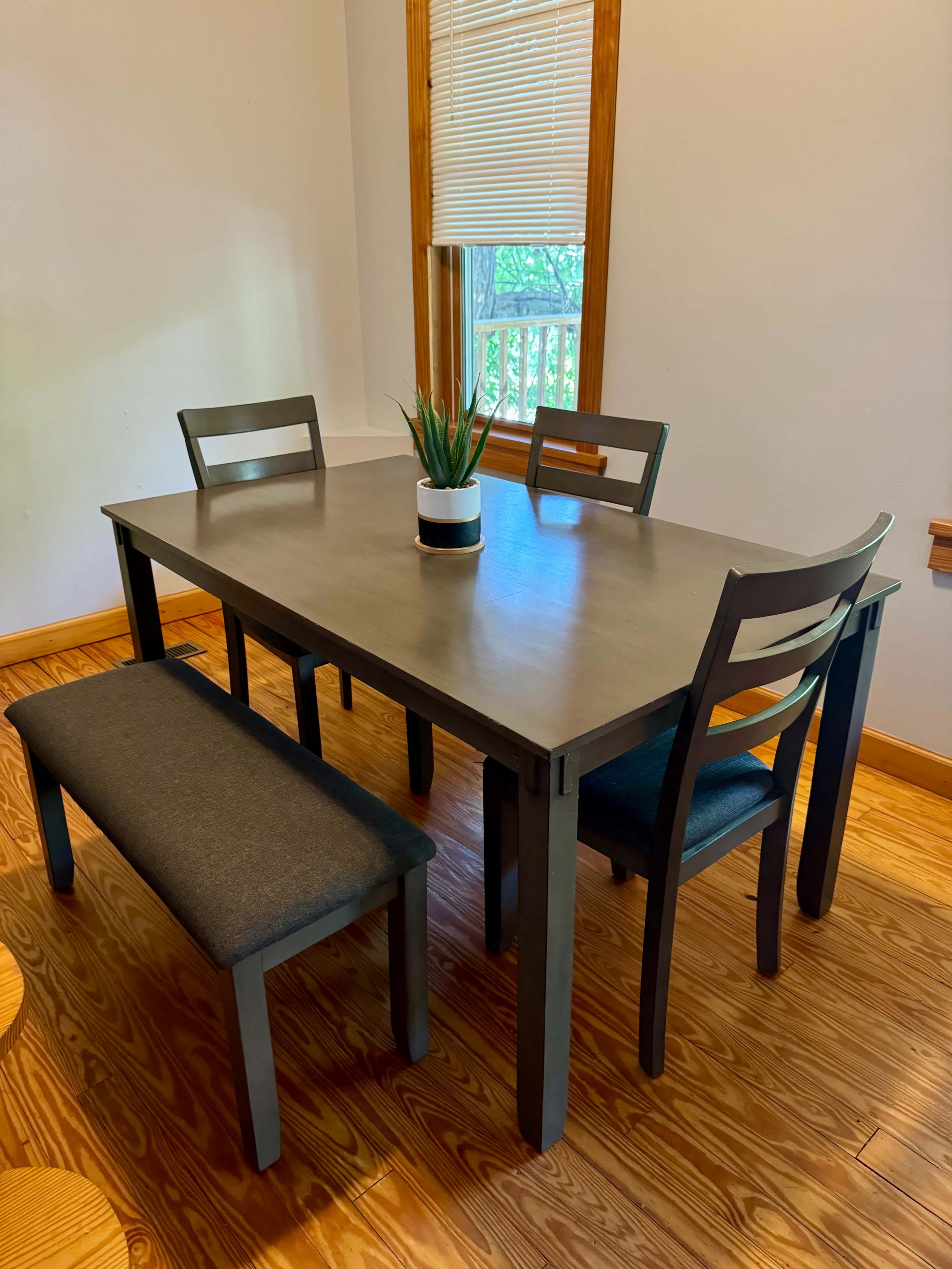 A wooden dining table with two chairs and a bench is positioned near a window in a room with wooden flooring.