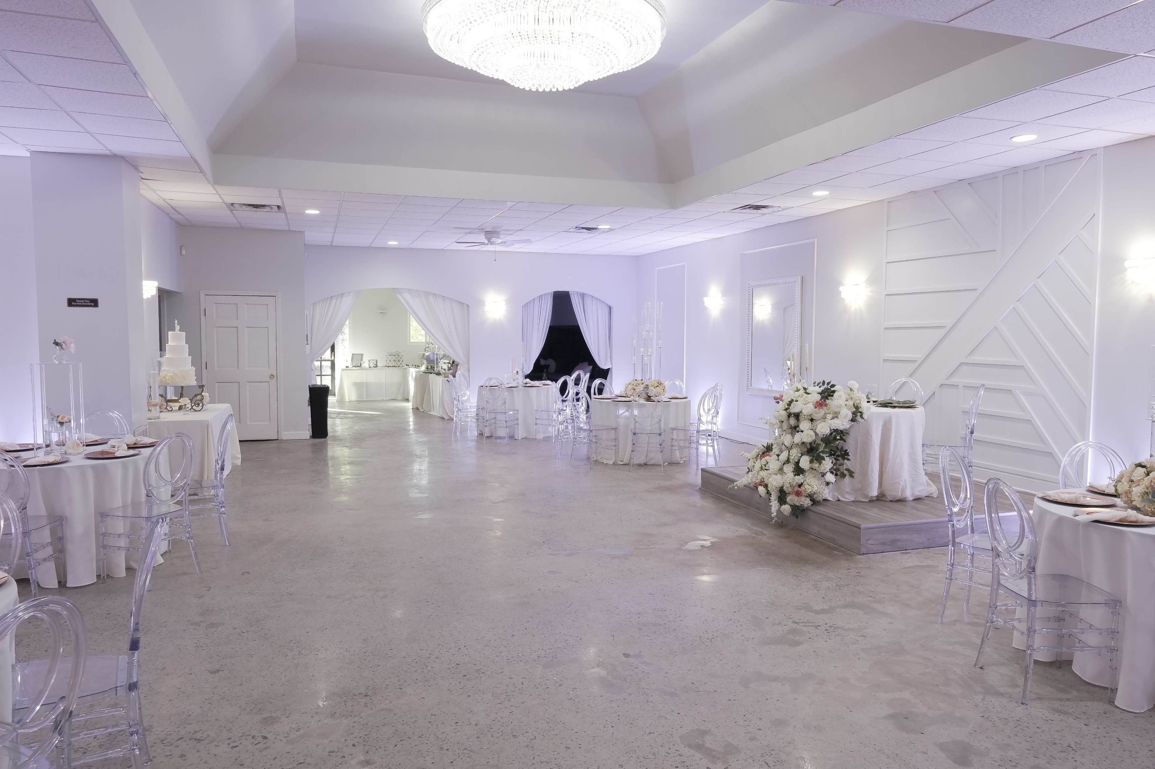 The image shows a spacious event hall set up for a banquet, featuring white tablecloths, clear chairs, and a decorated cake display.