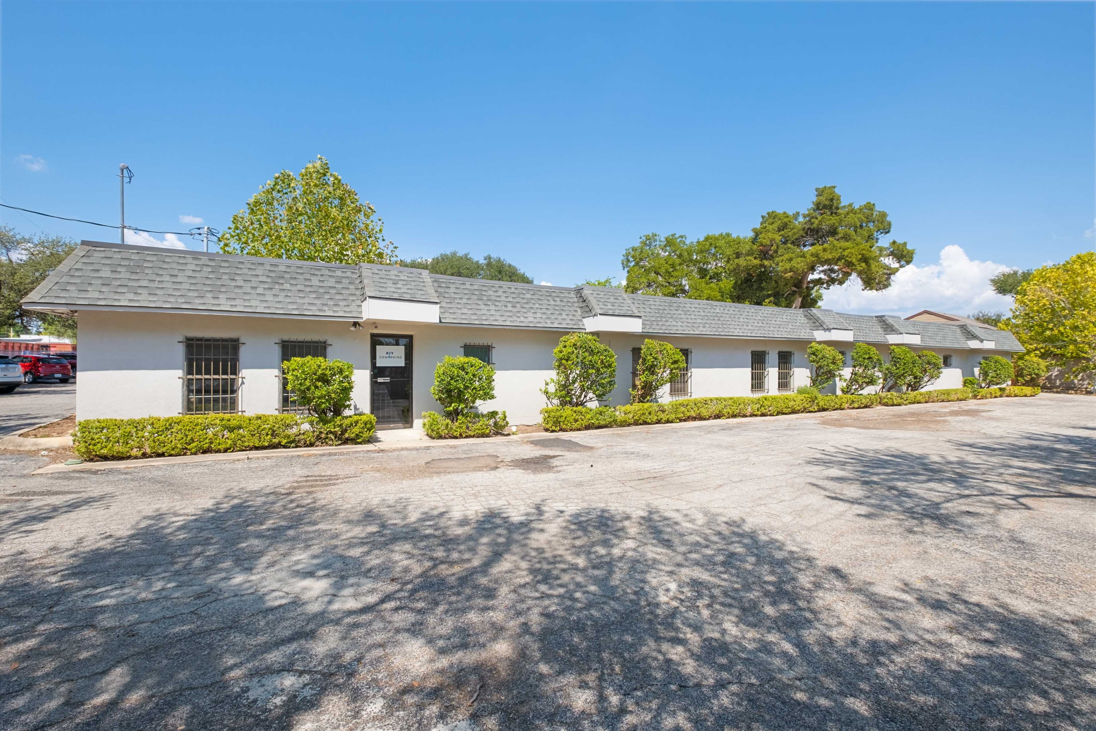 The image shows a single-story building with a gray roof and a row of evenly spaced windows, surrounded by manicured shrubs and trees under a clear blue sky.