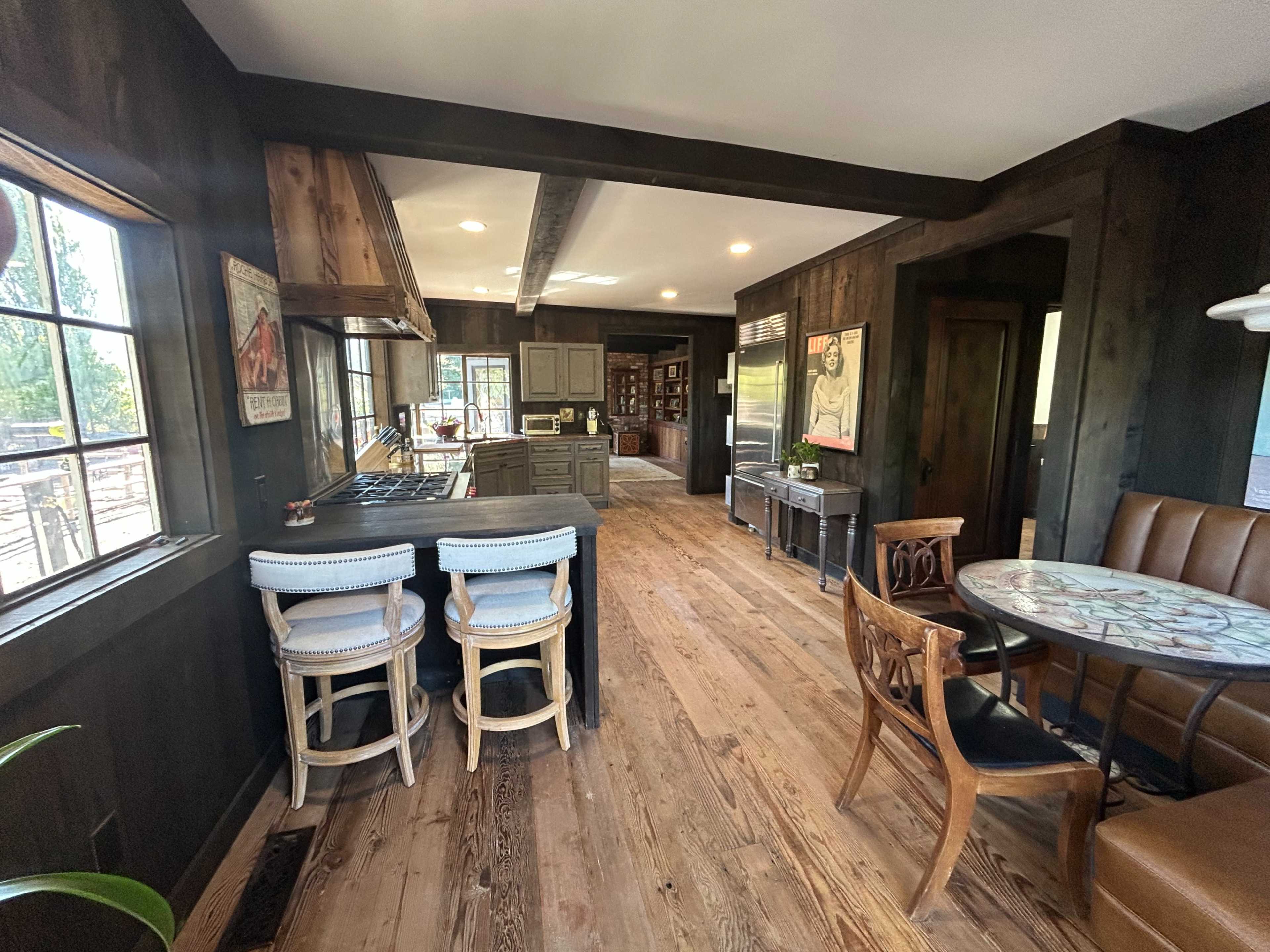 The image shows a rustic kitchen and dining area with wooden floors, dark walls, and large windows allowing natural light.