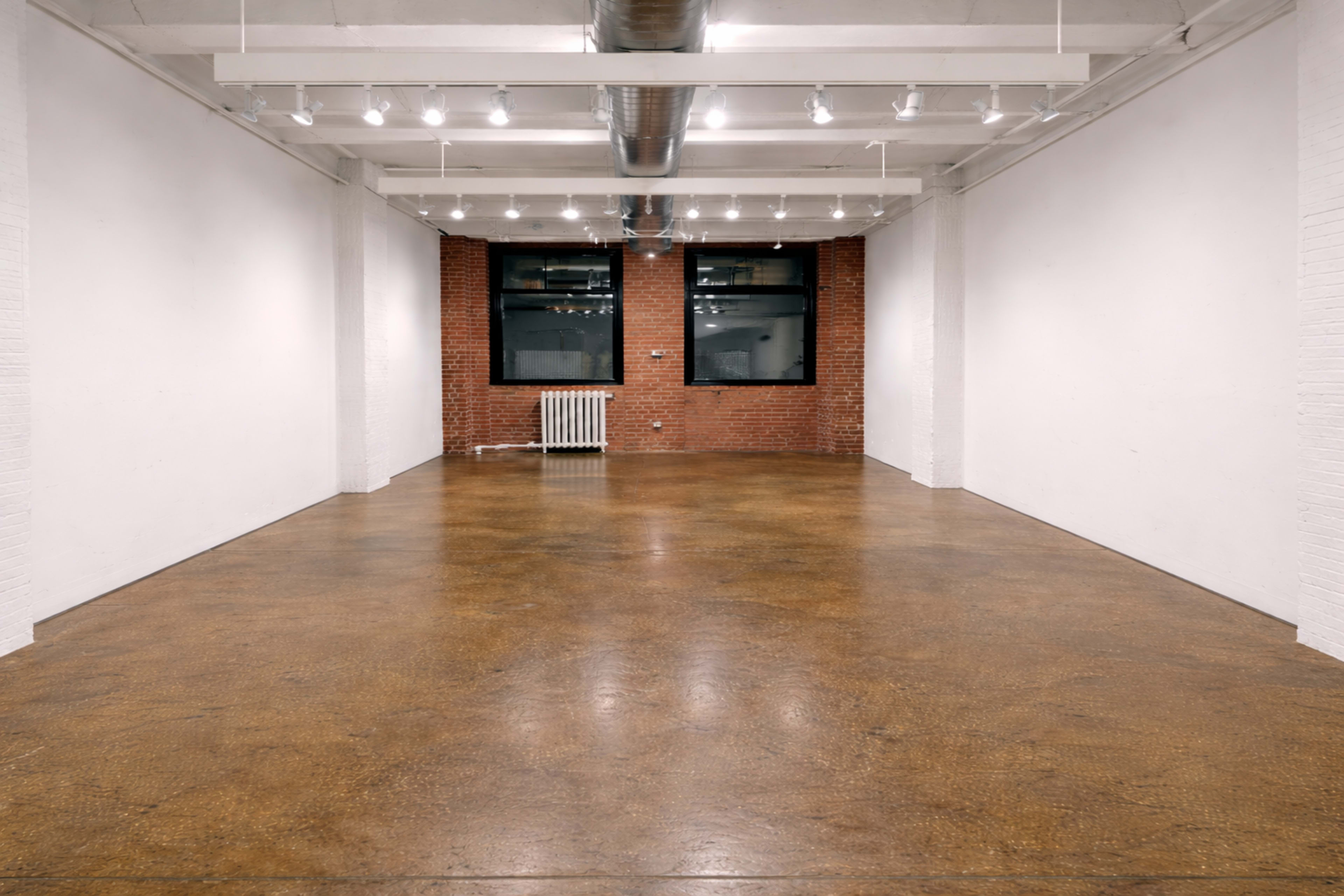 The image shows an empty, spacious room with polished concrete floors, exposed brick walls, and bright overhead lights.