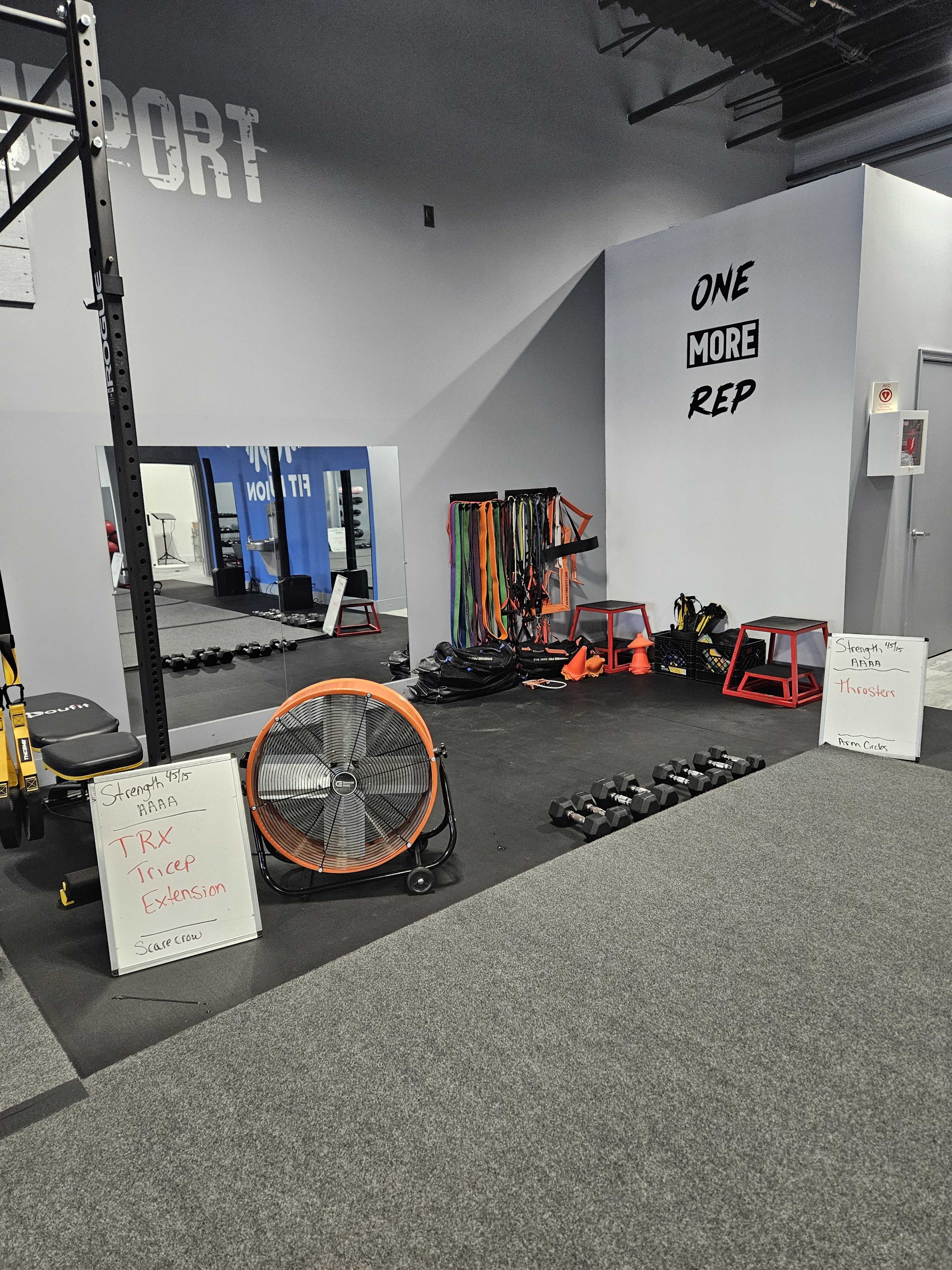 A gym corner with various workout equipment, including dumbbells, resistance bands, a fan, and workout schedules on whiteboards.