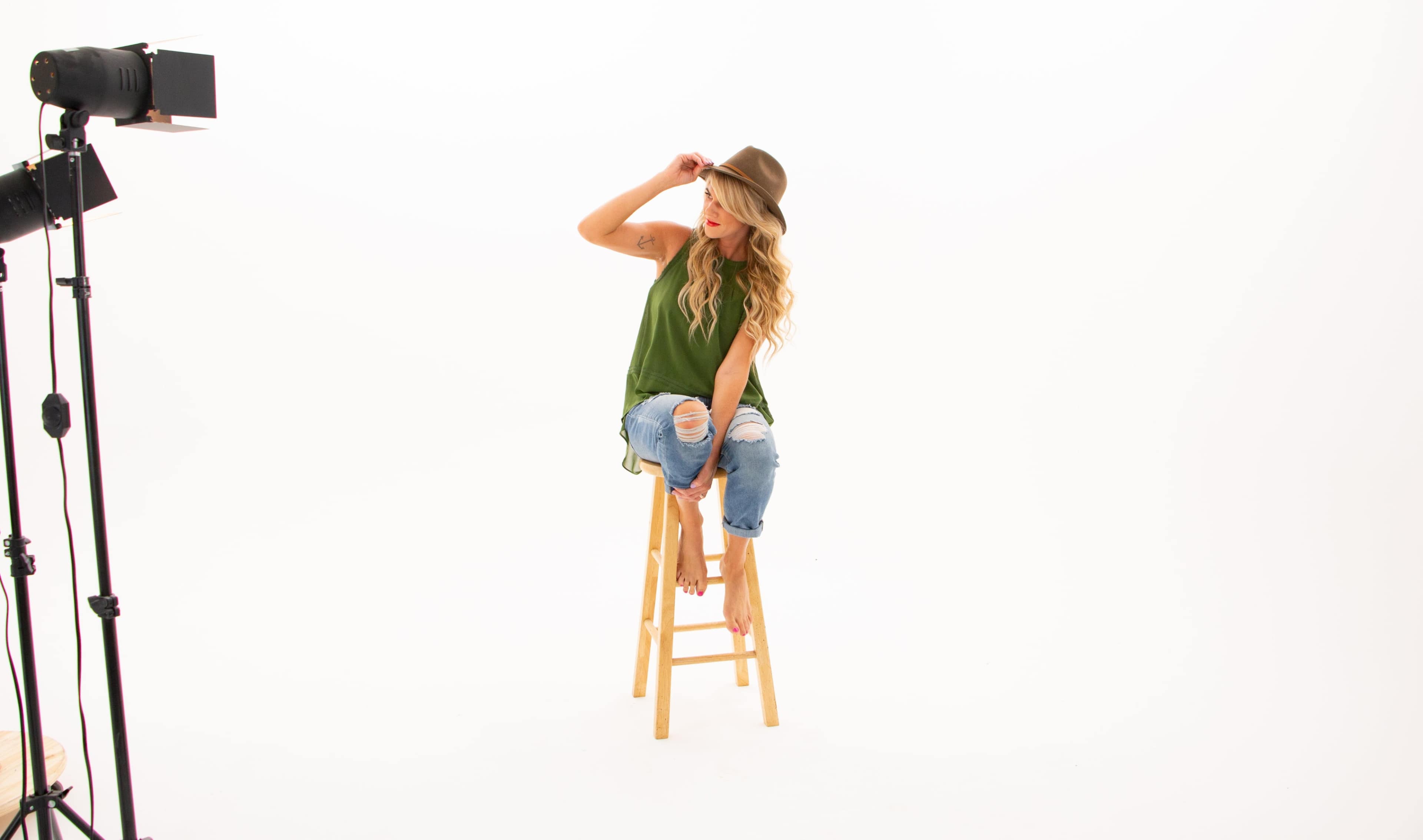 A woman wearing a green tank top and jeans sits on a wooden stool, adjusting a hat while facing the camera.