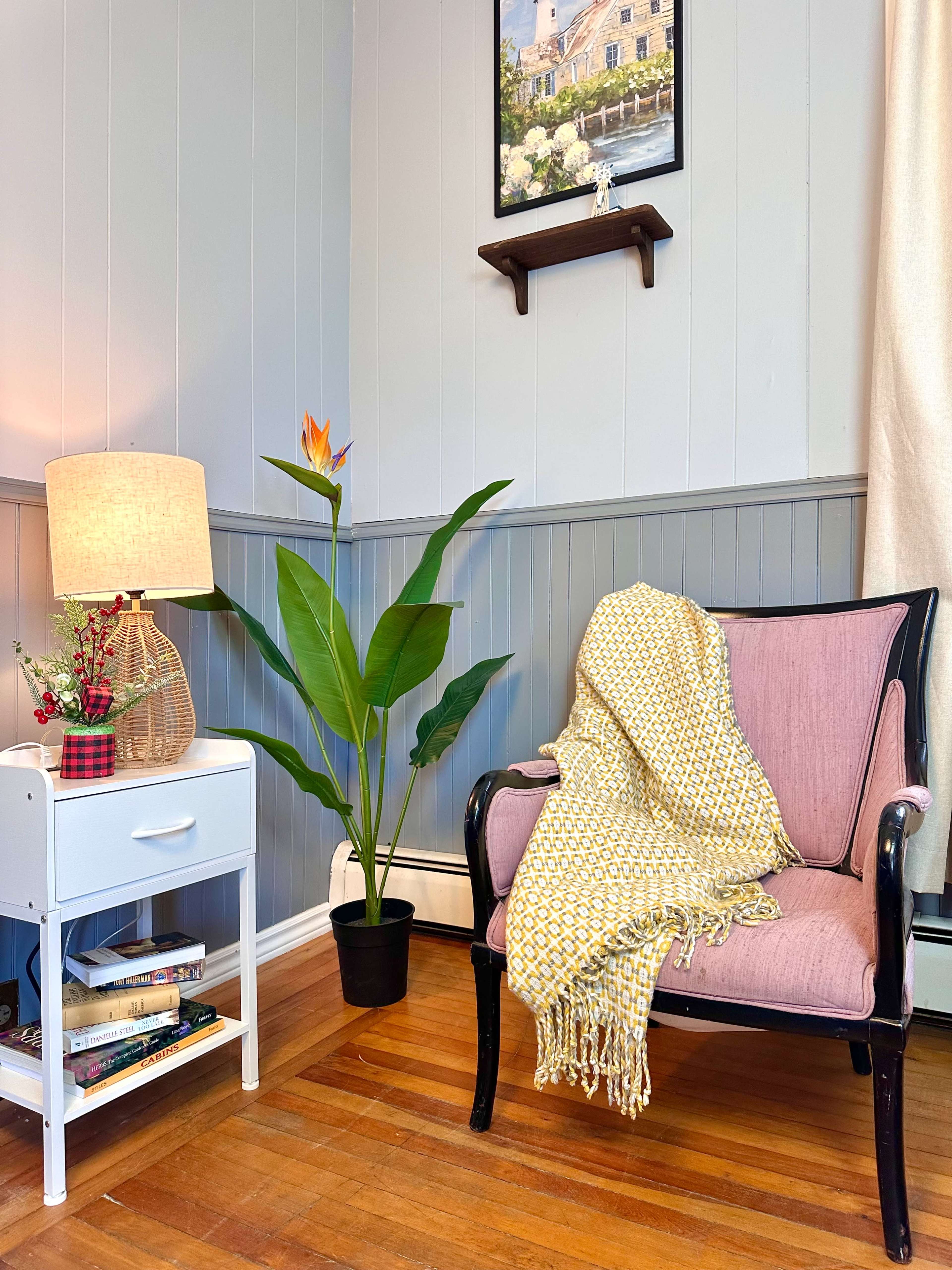 A pink chair with a yellow throw blanket sits beside a small white nightstand and a large green plant, with a lamp illuminating the corner of the room.