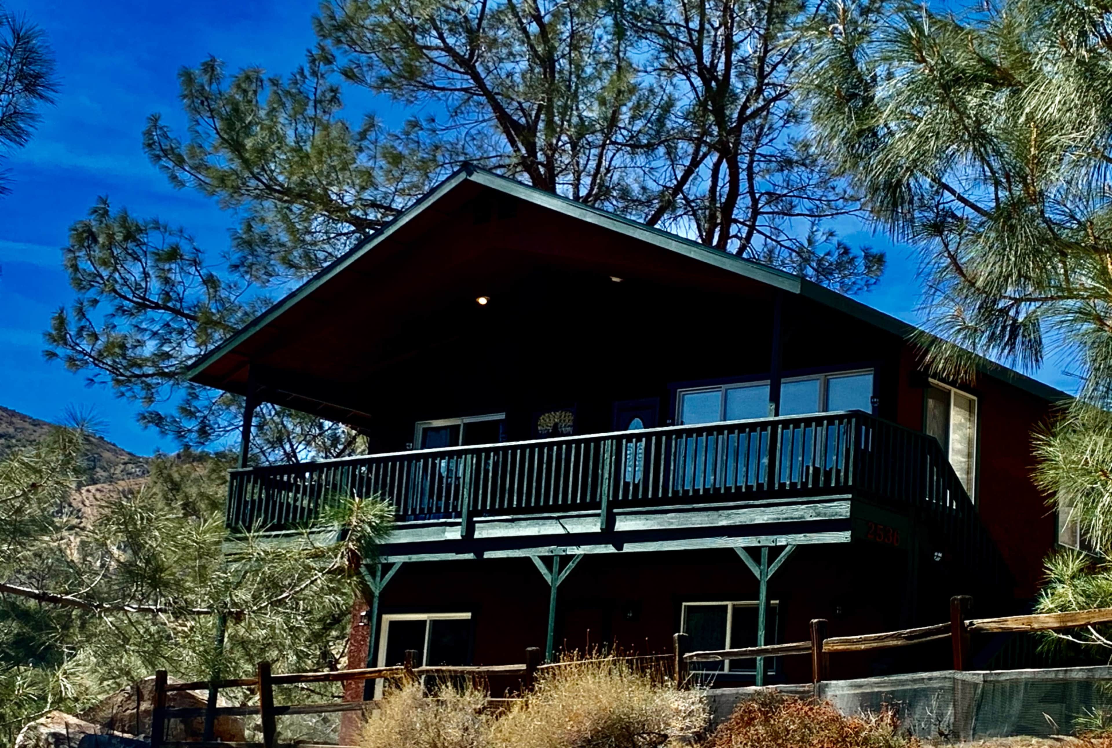 A red cabin with a black balcony is surrounded by pine trees against a blue sky.