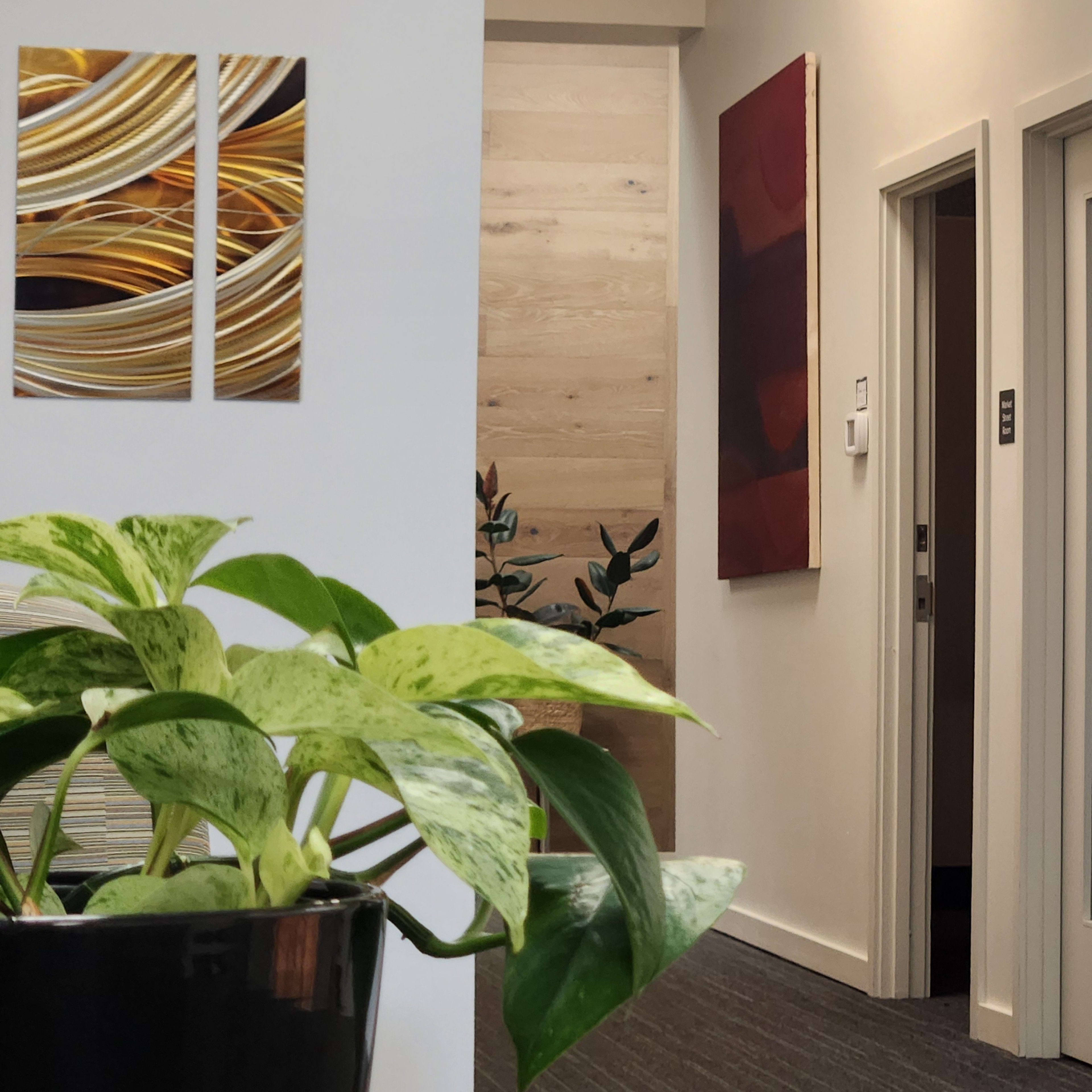 The image shows a modern interior with green plants in the foreground and a hallway leading to closed doors in the background, adorned with art on the walls.
