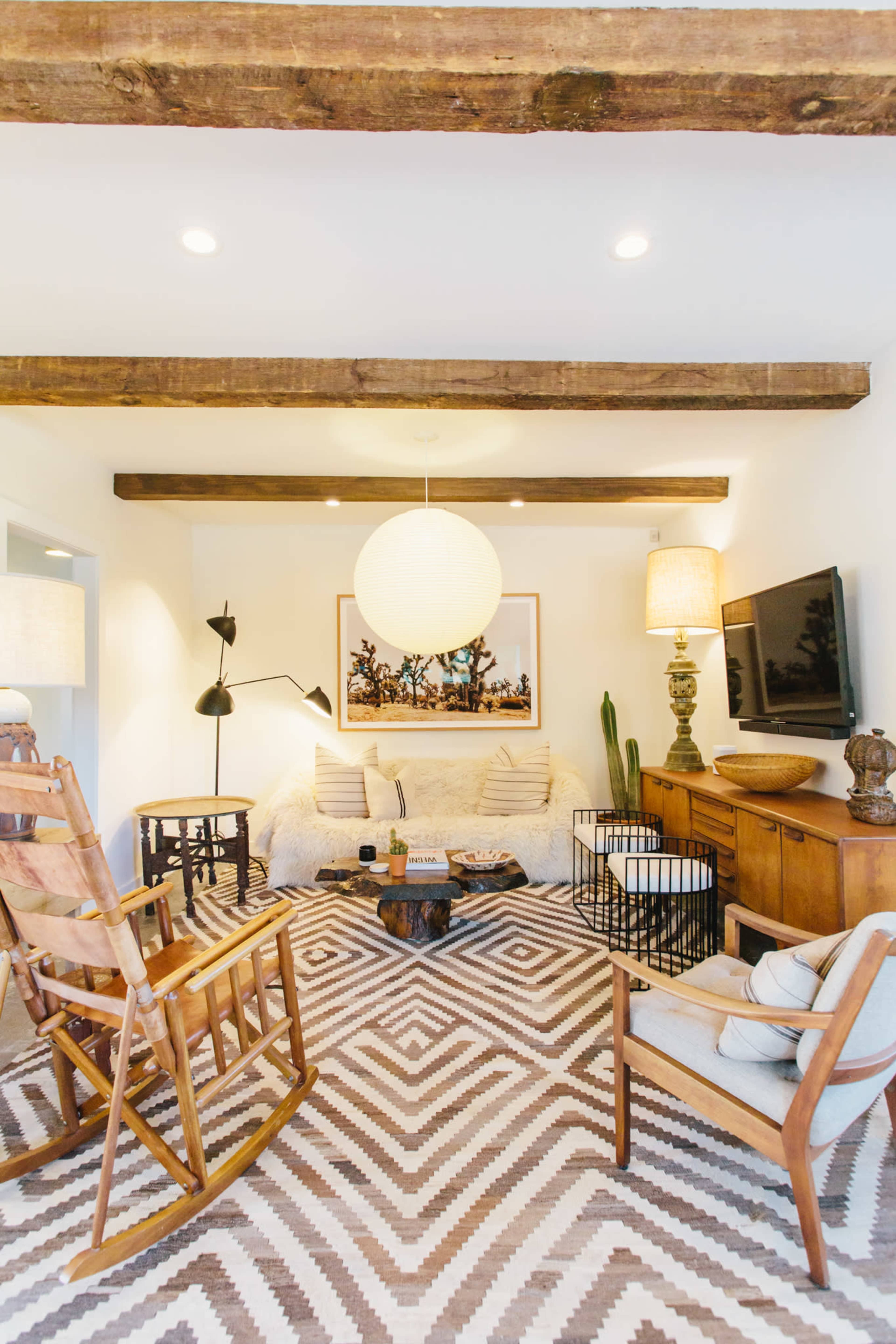 A cozy living room features a patterned rug, wooden beams, various seating options, and a large pendant light.