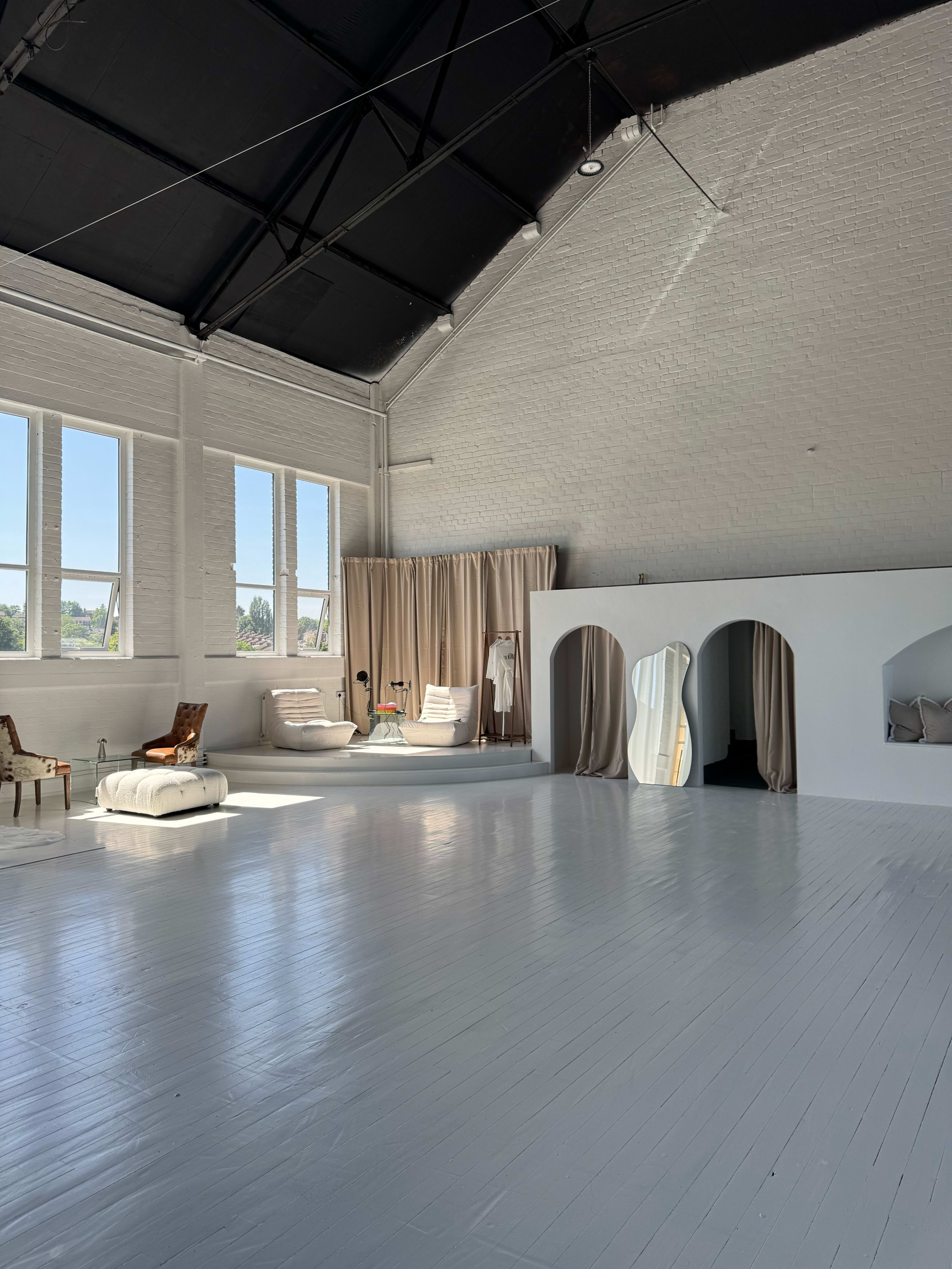 The image shows a spacious, bright interior with high ceilings, large windows, and minimalistic furniture arranged against white walls.
